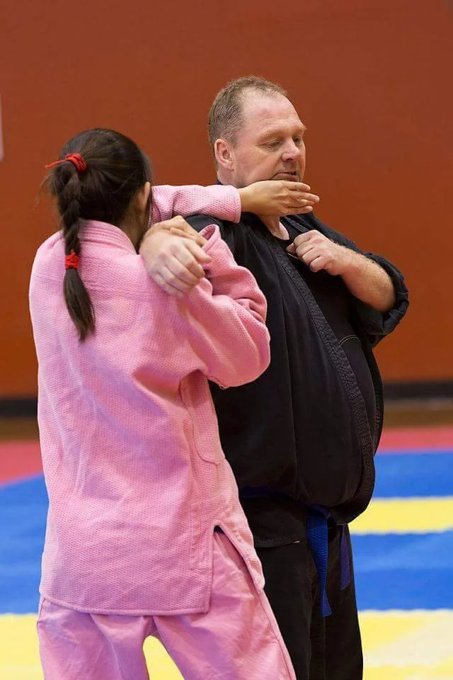 A girl in pink martial arts uniform practicing a self-defense move on an instructor in a dojo with colorful mats.