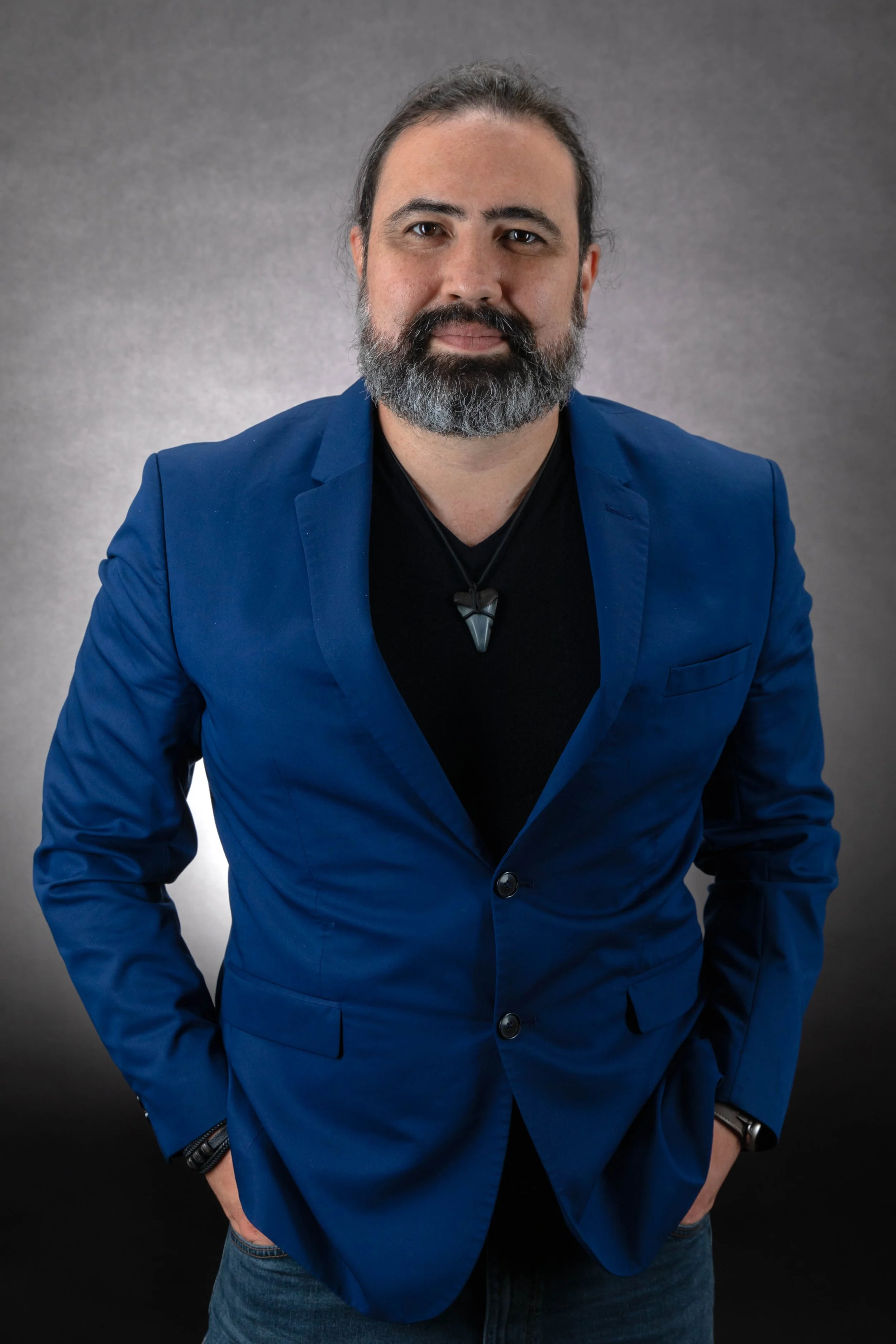 Professional photo of Danny Bravo with dark hair and a beard, wearing a blue blazer, black shirt, and a necklace, standing against a grey background.