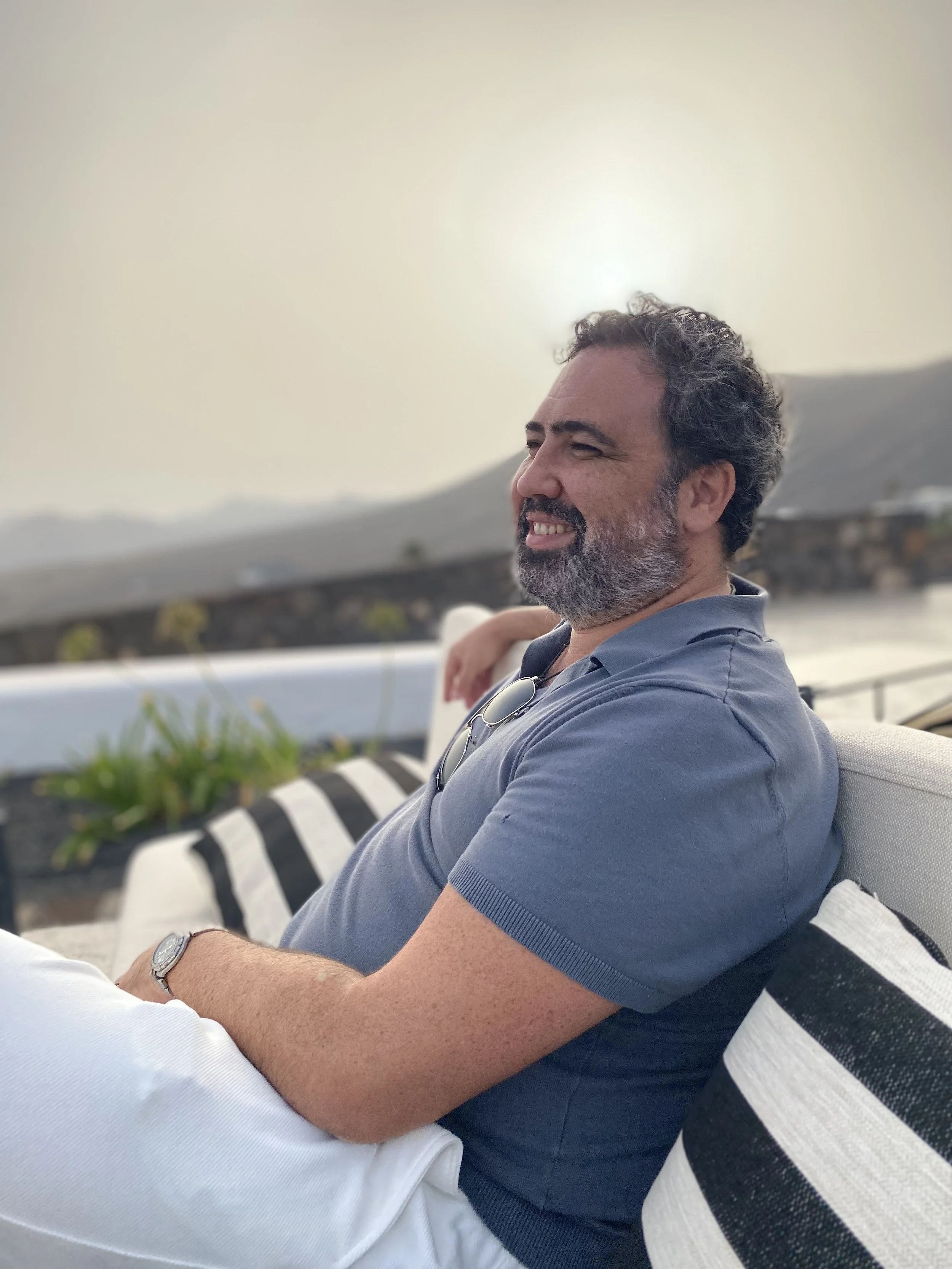 Danny sitting outdoors on a bench, smiling and looking to the side, with mountains and a pale sky in the background.