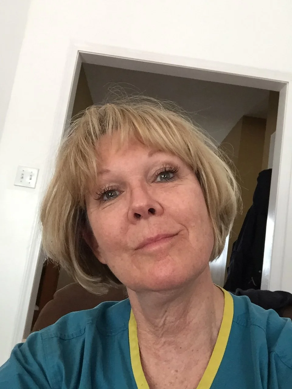A woman with short blonde hair and blue eyes taking a selfie indoors, wearing a blue shirt with yellow trim.