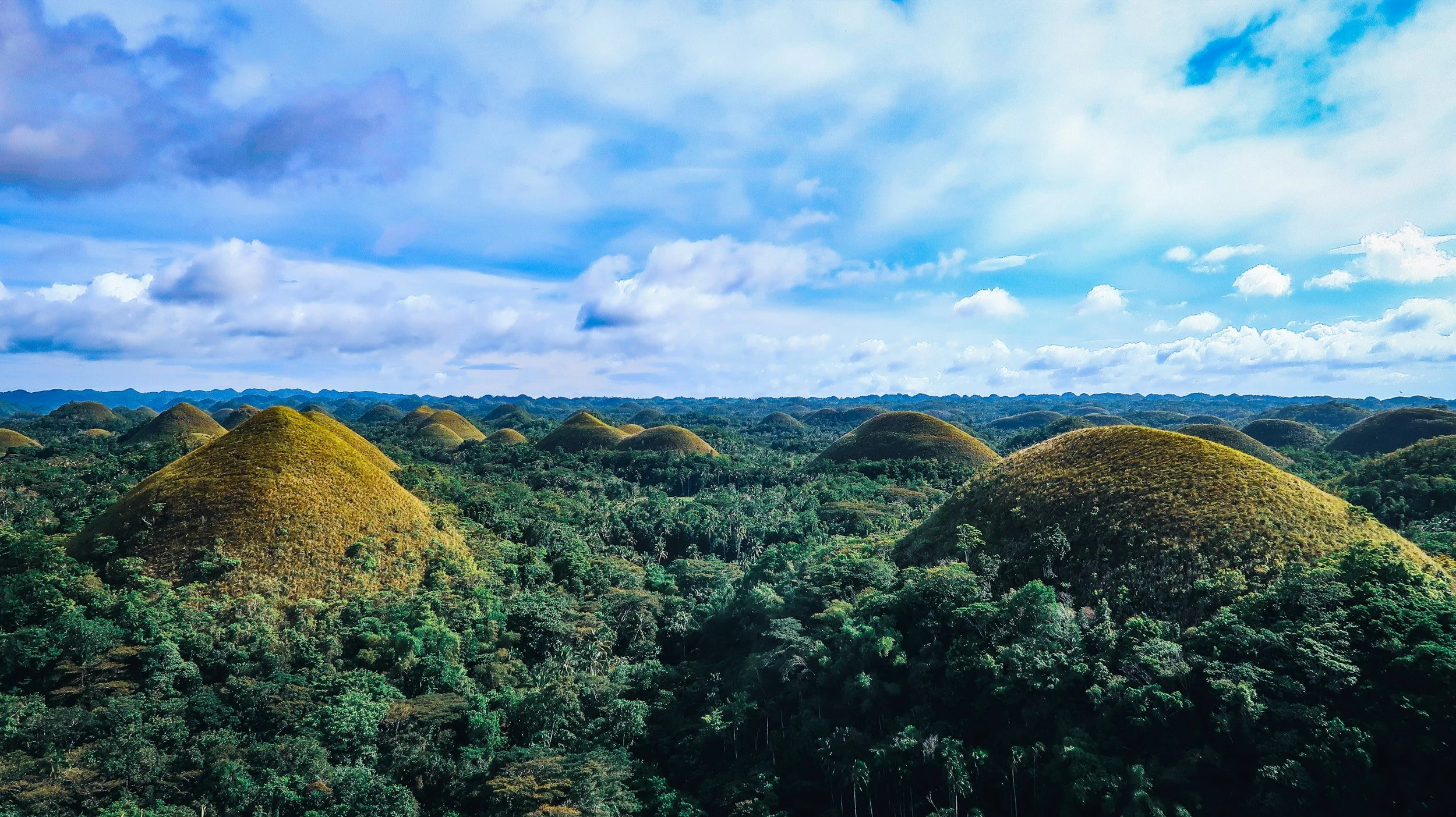 Landscape view of the Chocolate Hills in a lush green landscape under a partly cloudy sky.