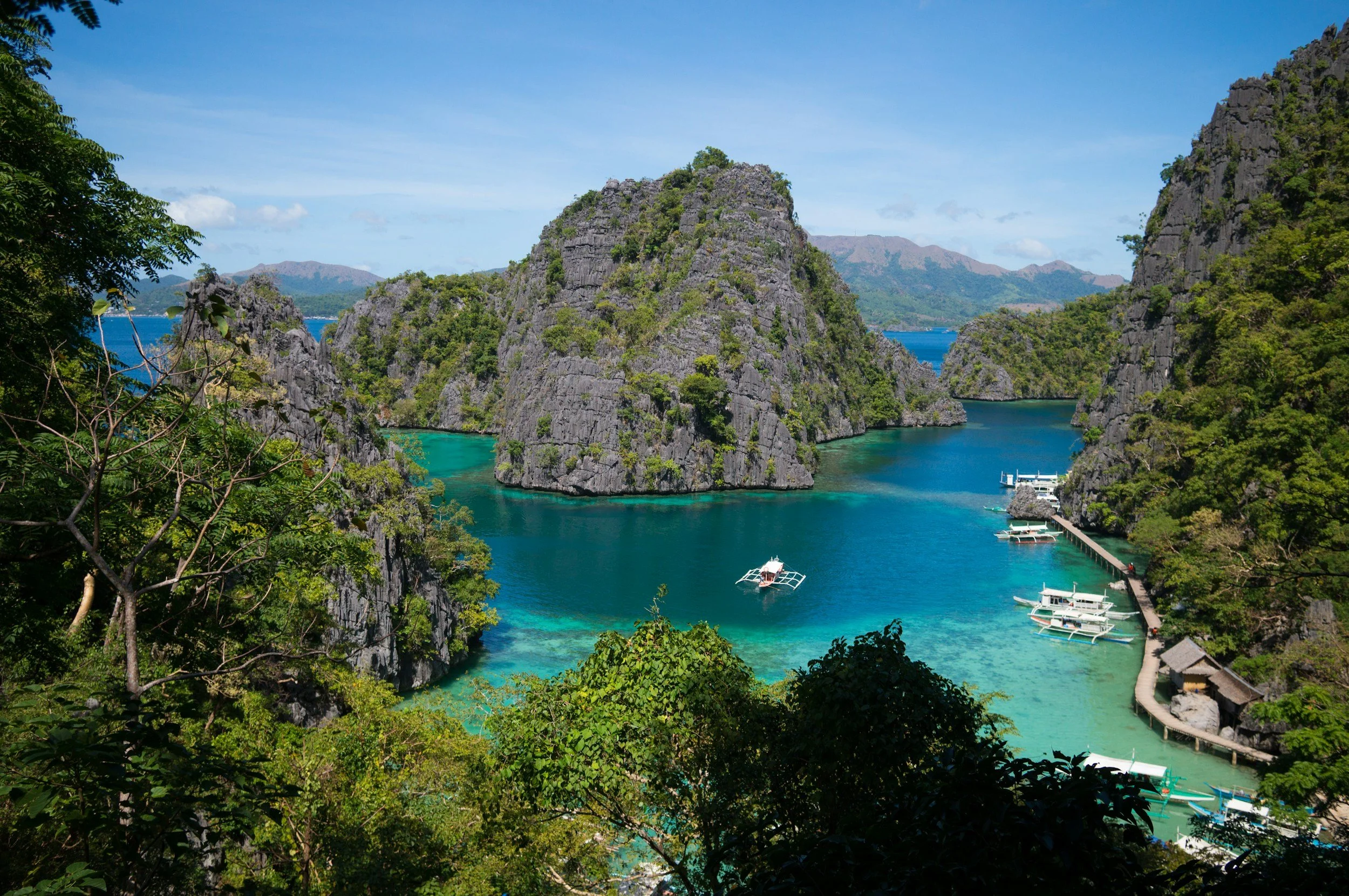 Tropical island landscape with turquoise water, rocky cliffs covered in green vegetation, and boats docked along a pier, under a blue sky.