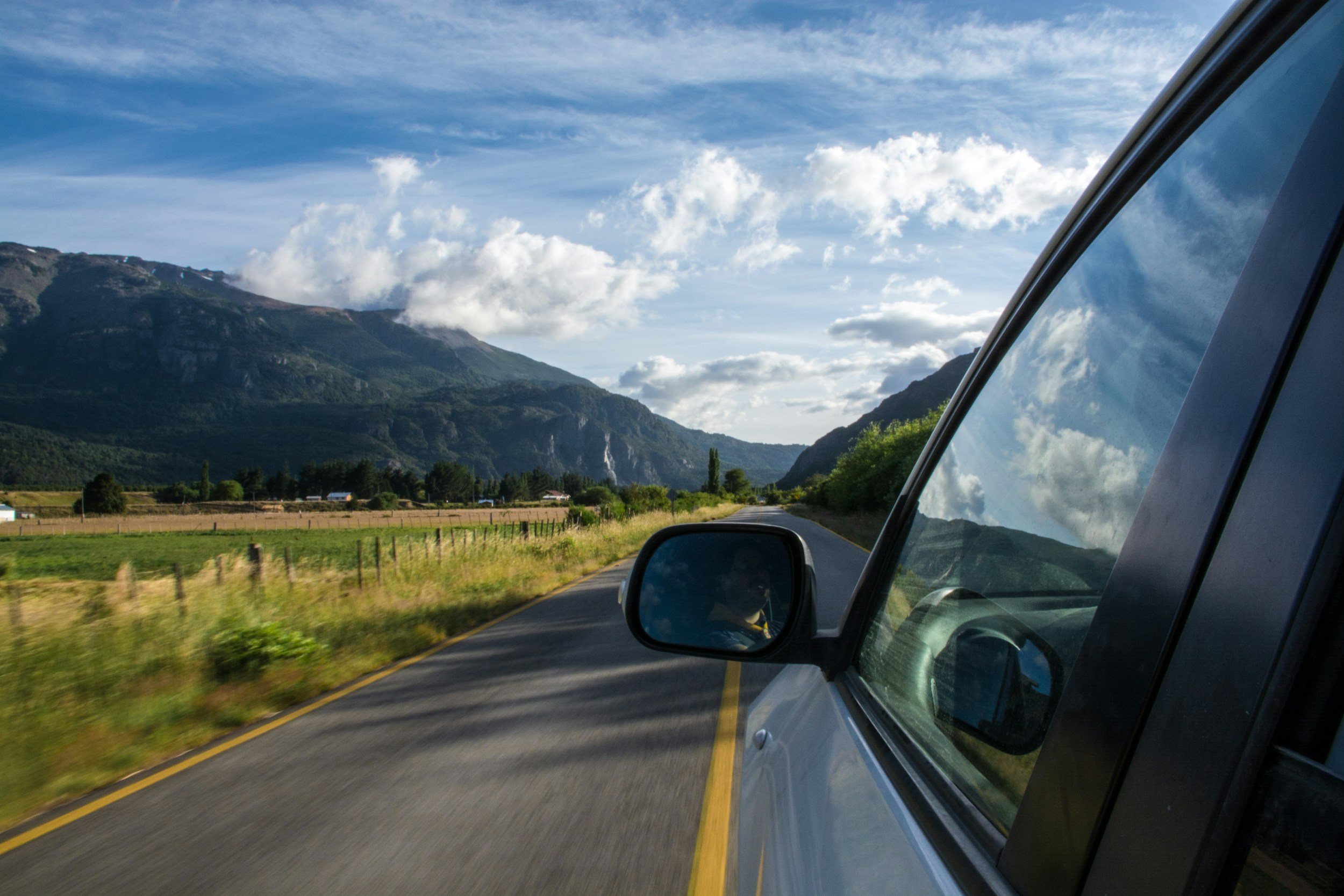 View from a car window on a rural road with mountains in the distance and a partly cloudy sky.