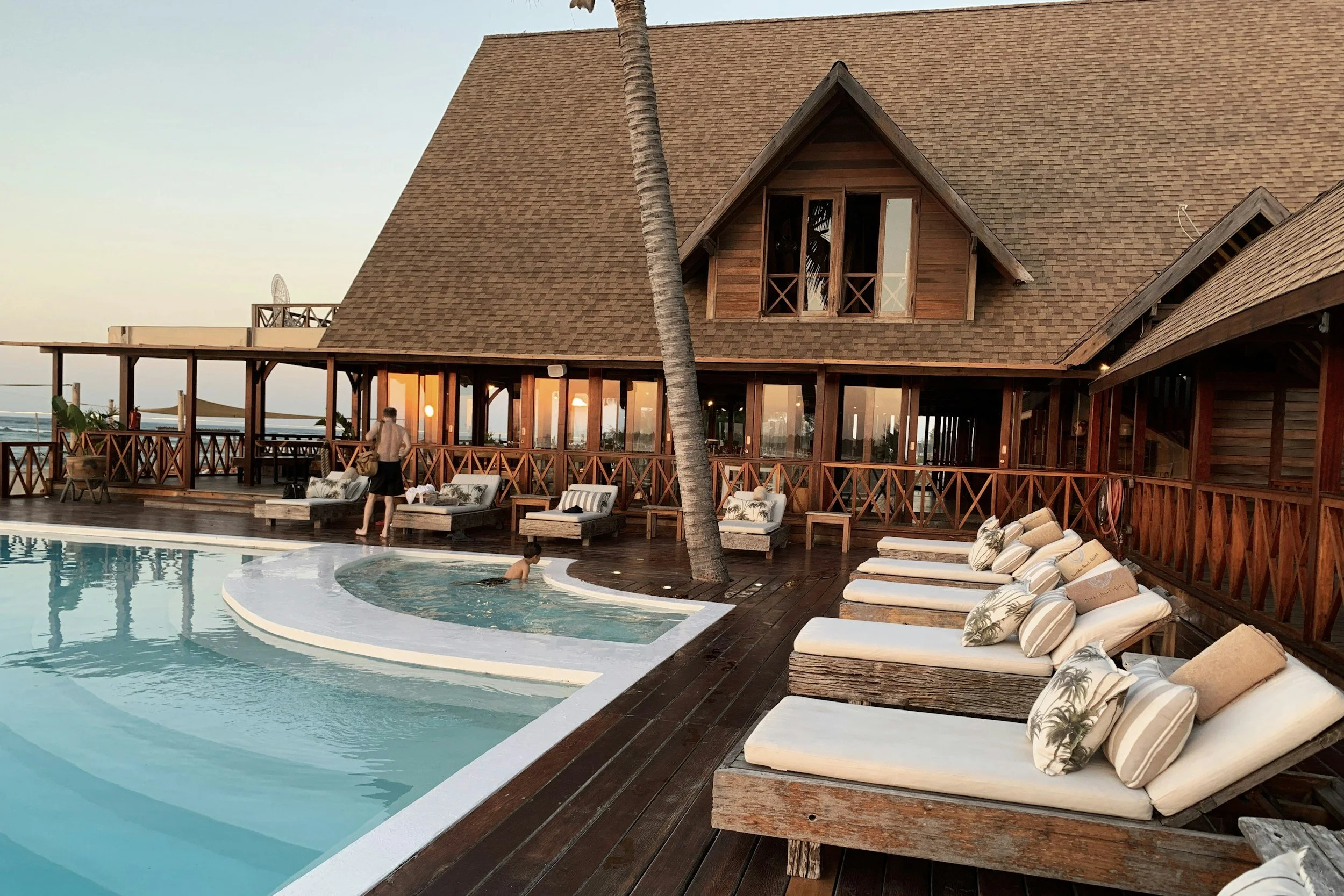 A luxurious wooden hotel with a poolside deck, lounge chairs, and a palm tree in the foreground, during sunset.