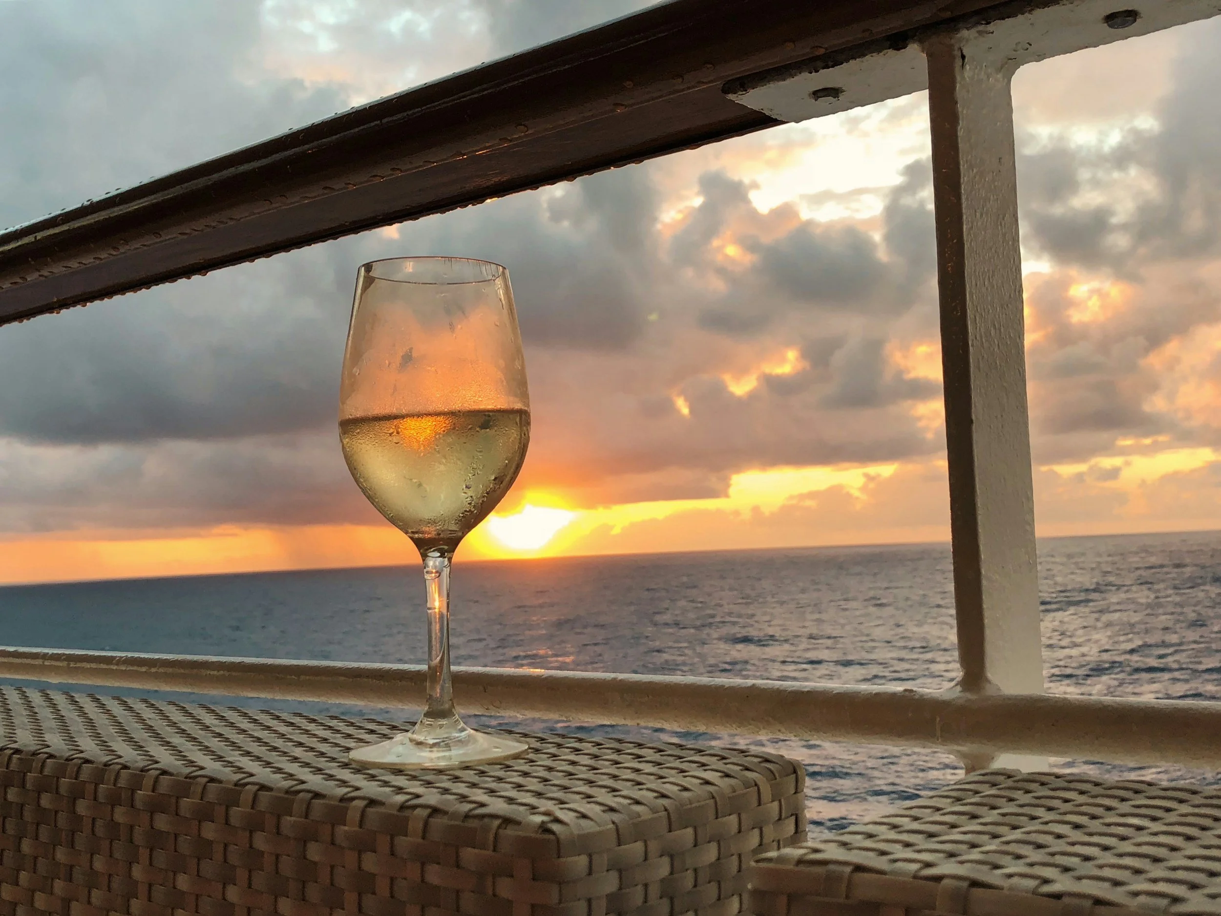 A glass of white wine on a woven table with a sunset view over the ocean in the background.