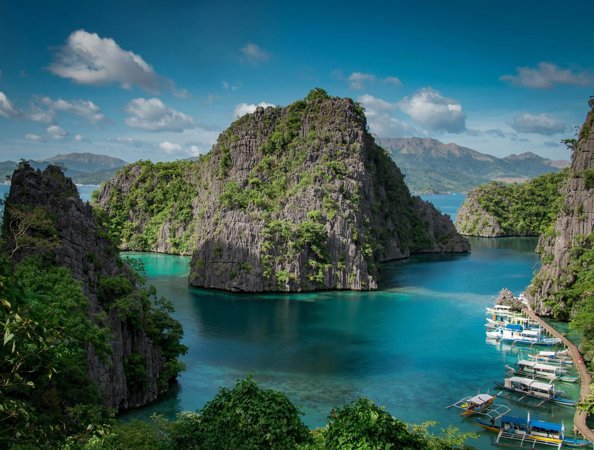 Tropical bays surrounded by tall, green, rocky cliffs, with docked boats along a narrow pier on a sunny day with a partly cloudy sky.