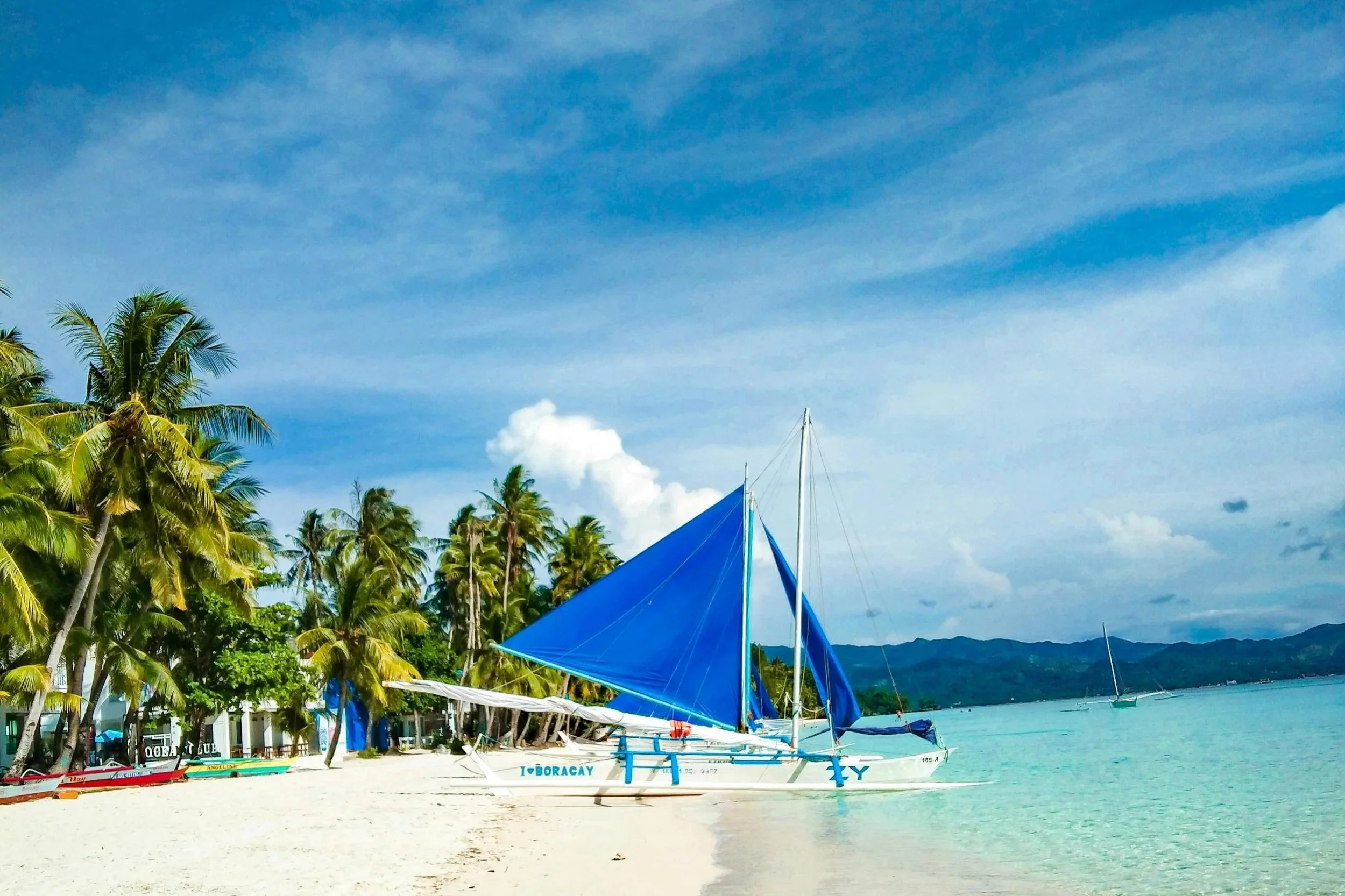 A sailboat with a blue sail anchored on a sandy beach with palm trees and a blue ocean under a partly cloudy sky.