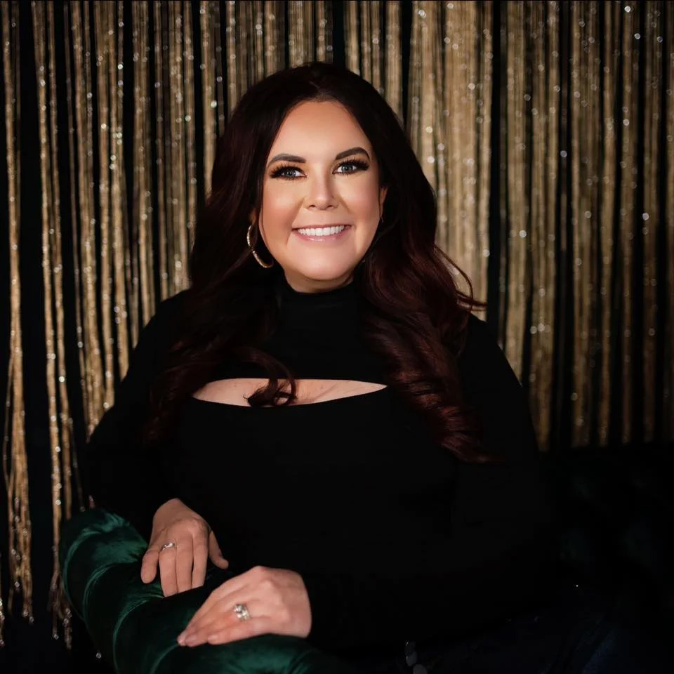 A woman with long dark hair and earrings, smiling, wearing a black top with a cutout detail, sitting in front of a gold glittery backdrop.