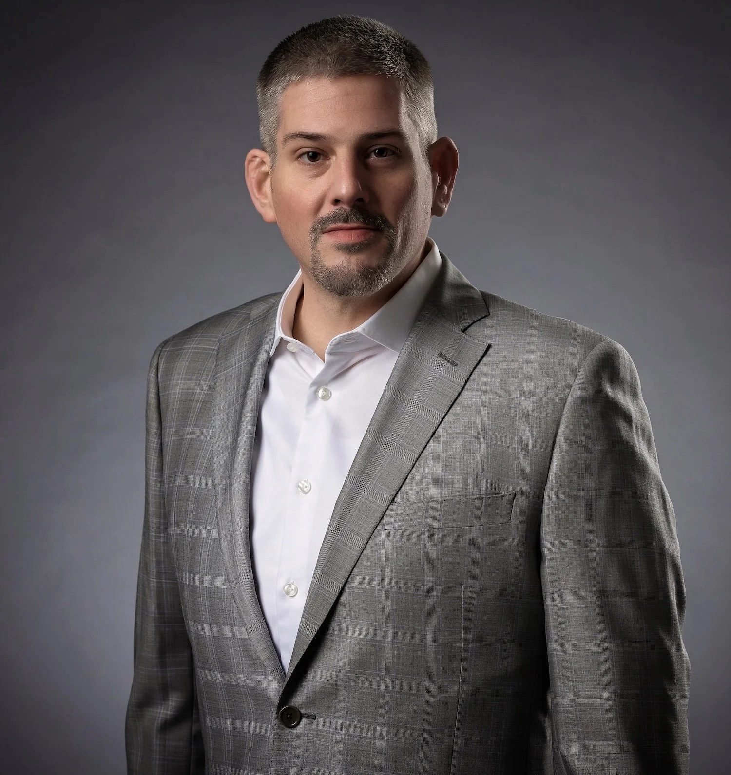 A man in a light gray suit with a white shirt, standing against a gray background.