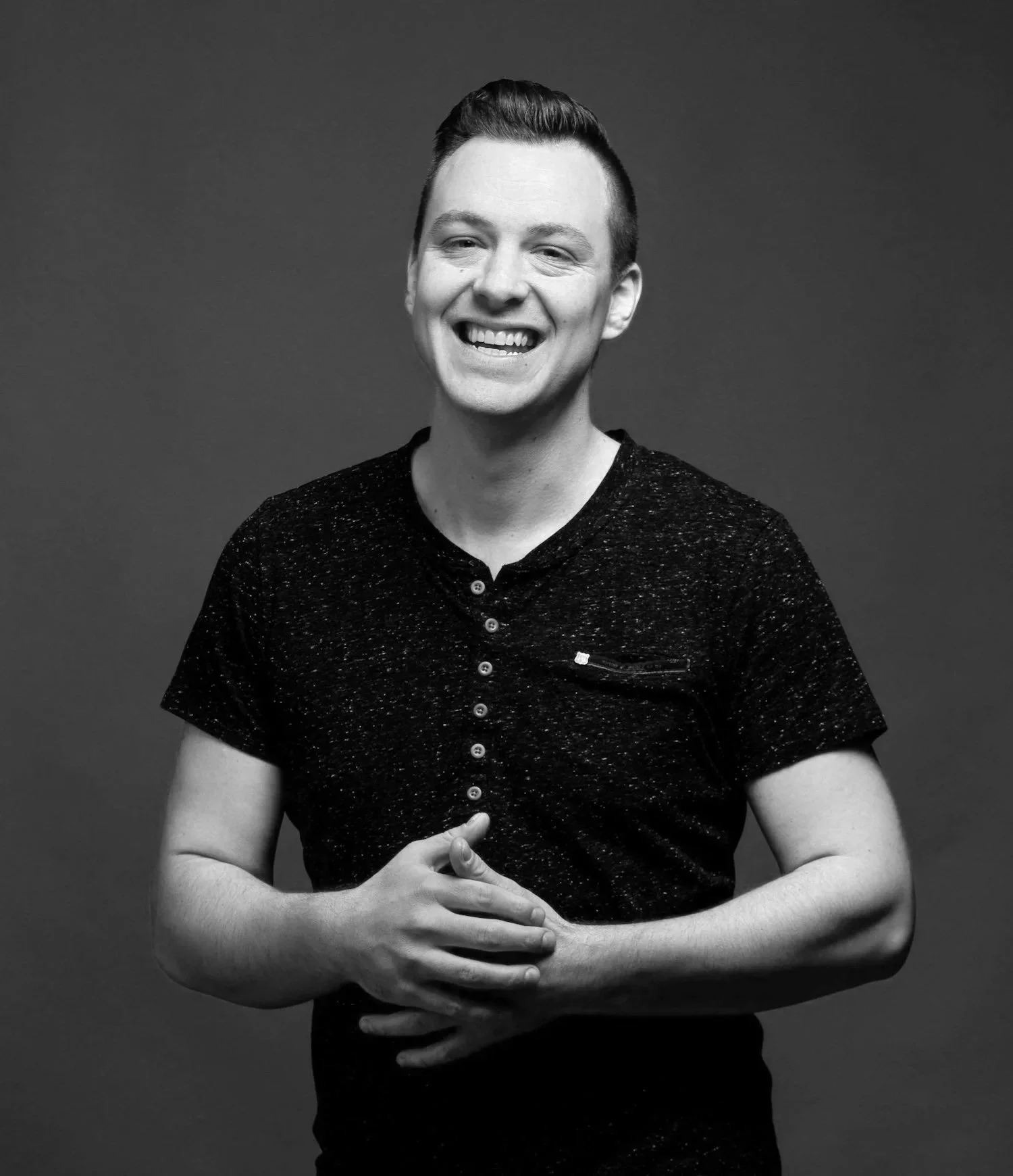 Black and white photo of smiling young man with short hair, wearing a dark speckled t-shirt, standing against a gray background.