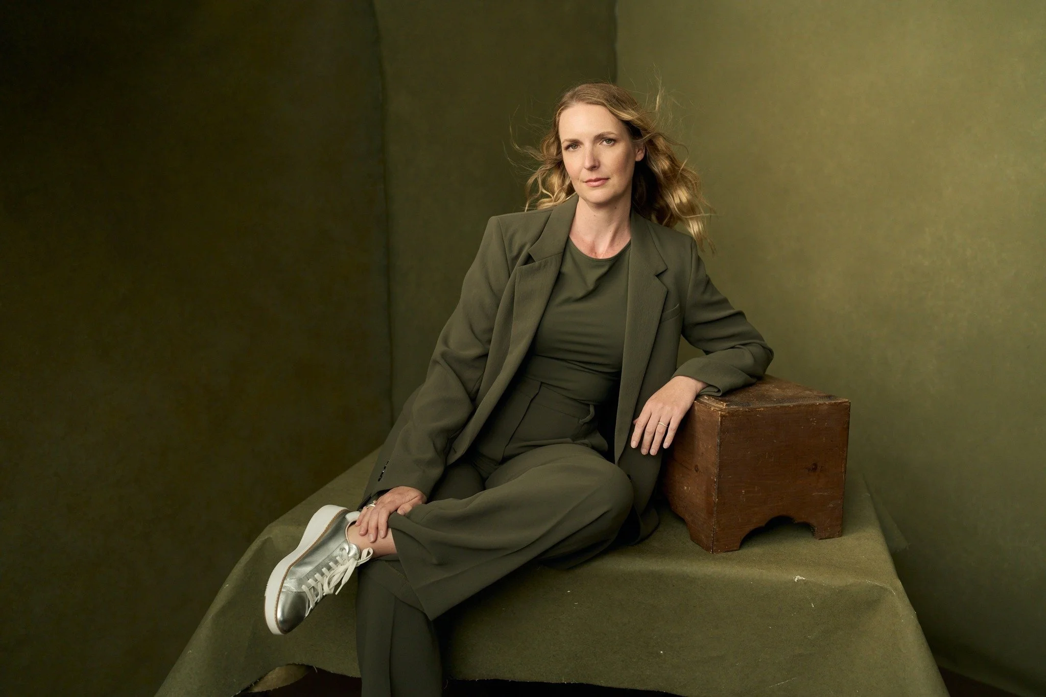 A woman with shoulder-length curly brown hair and hazel eyes looks at the camera, standing against a yellow wall, wearing a black top.