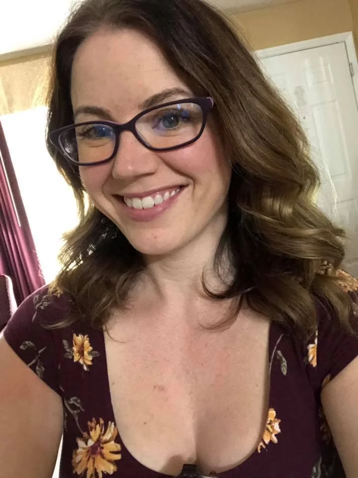 A woman with curly brown hair, wearing glasses and a floral dress, smiling for the camera indoors.