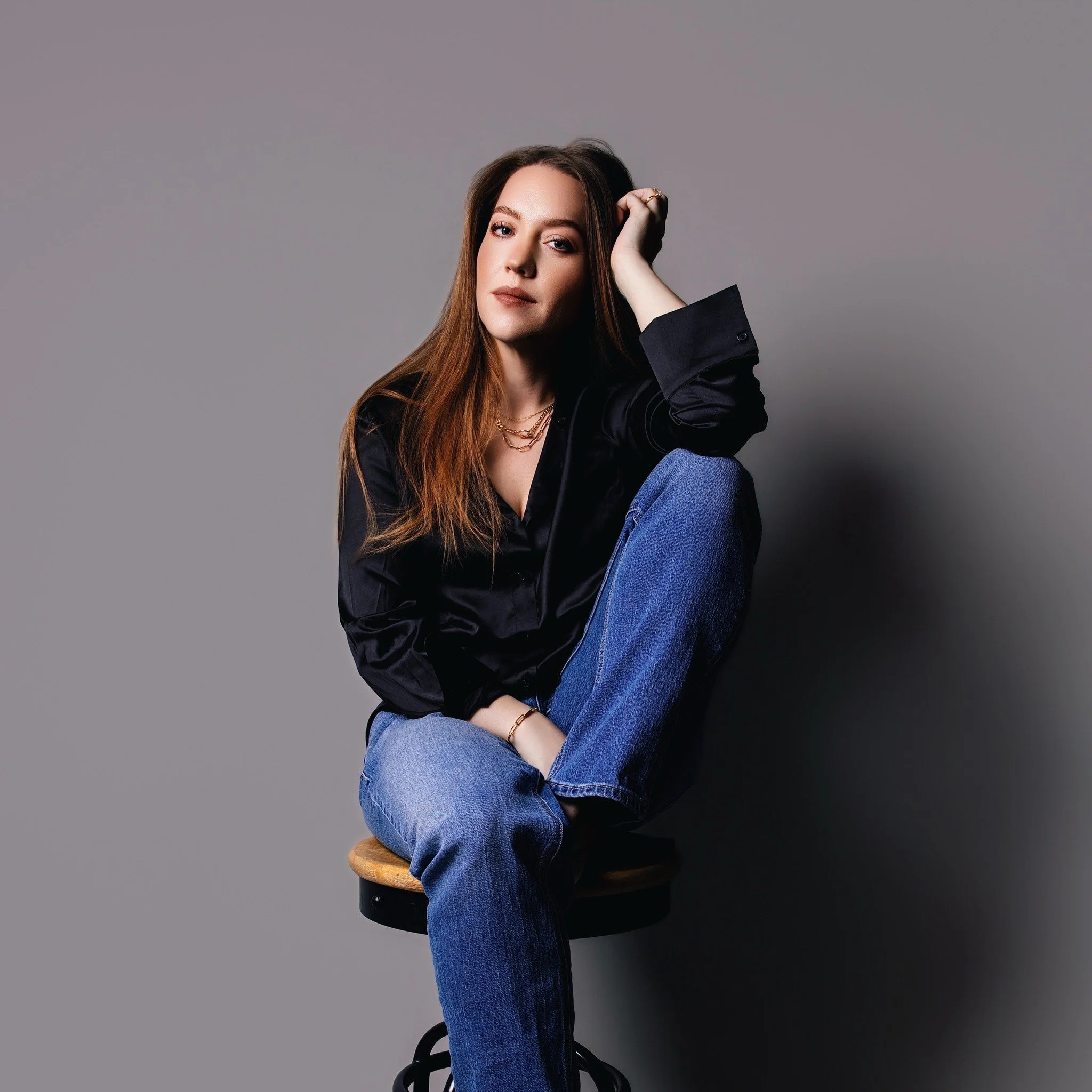 Young woman with long brown hair sitting on a wooden stool against a gray background, wearing a black satin shirt and blue jeans.