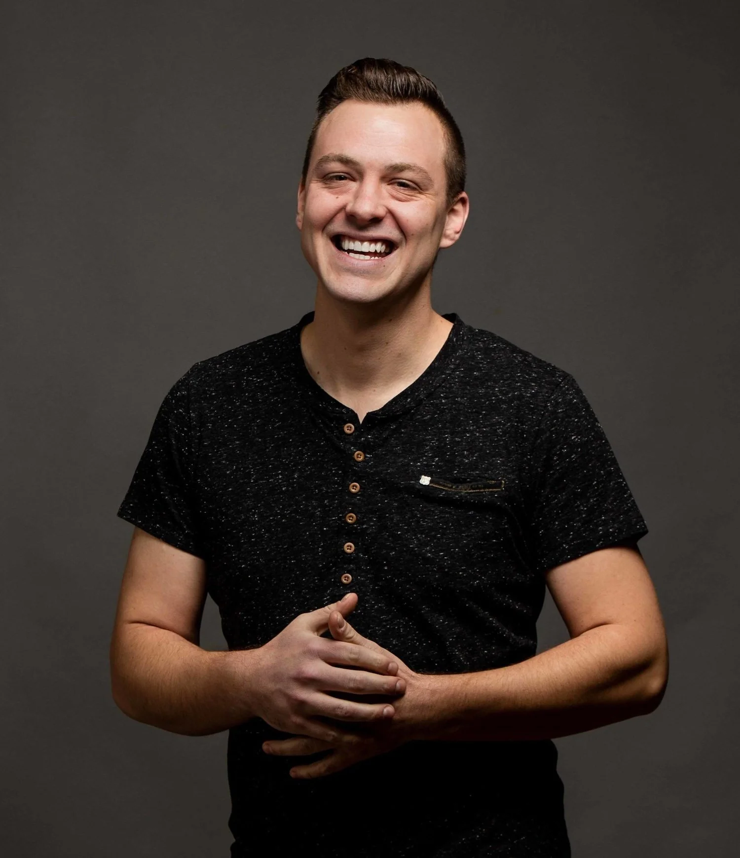 Smiling young man with short hair, wearing a black speckled t-shirt, standing against a dark gray background.