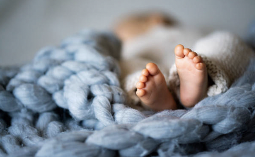 Close-up of baby feet peeking out from under a knitted blanket, surrounded by a soft, chunky gray blanket.