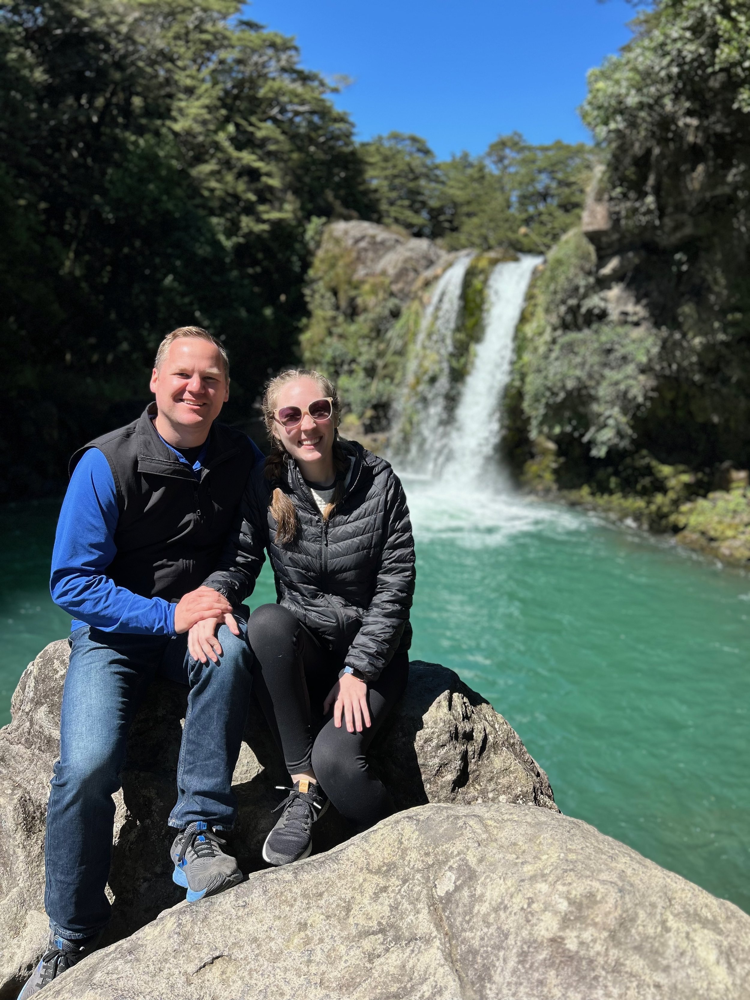 A smiling couple sitting on a large rock in front of a waterfall with lush green trees and a clear blue sky in the background.