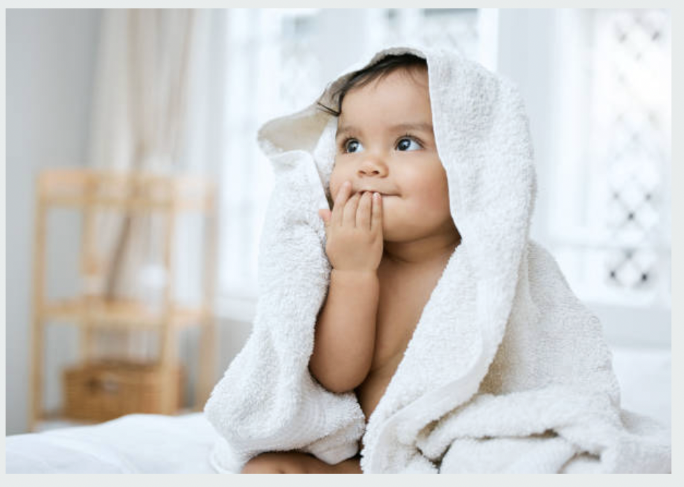 A toddler with big blue eyes sitting on a bed wrapped in a white bath towel, with one hand near her mouth, in a bright room with a blurred background.
