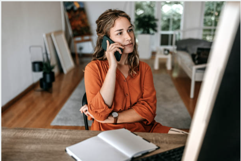 A woman with wavy blonde hair, wearing an orange blouse, is sitting at a desk in a bright room with large windows. She is talking on the phone and looking at her computer monitor with a notebook open in front of her.
