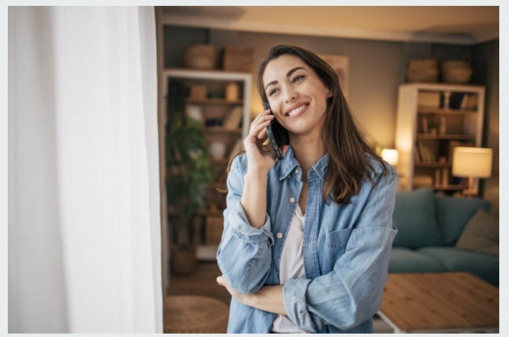 A woman in a denim shirt talking on the phone inside a cozy living room with bookshelves, plants, and a sofa.