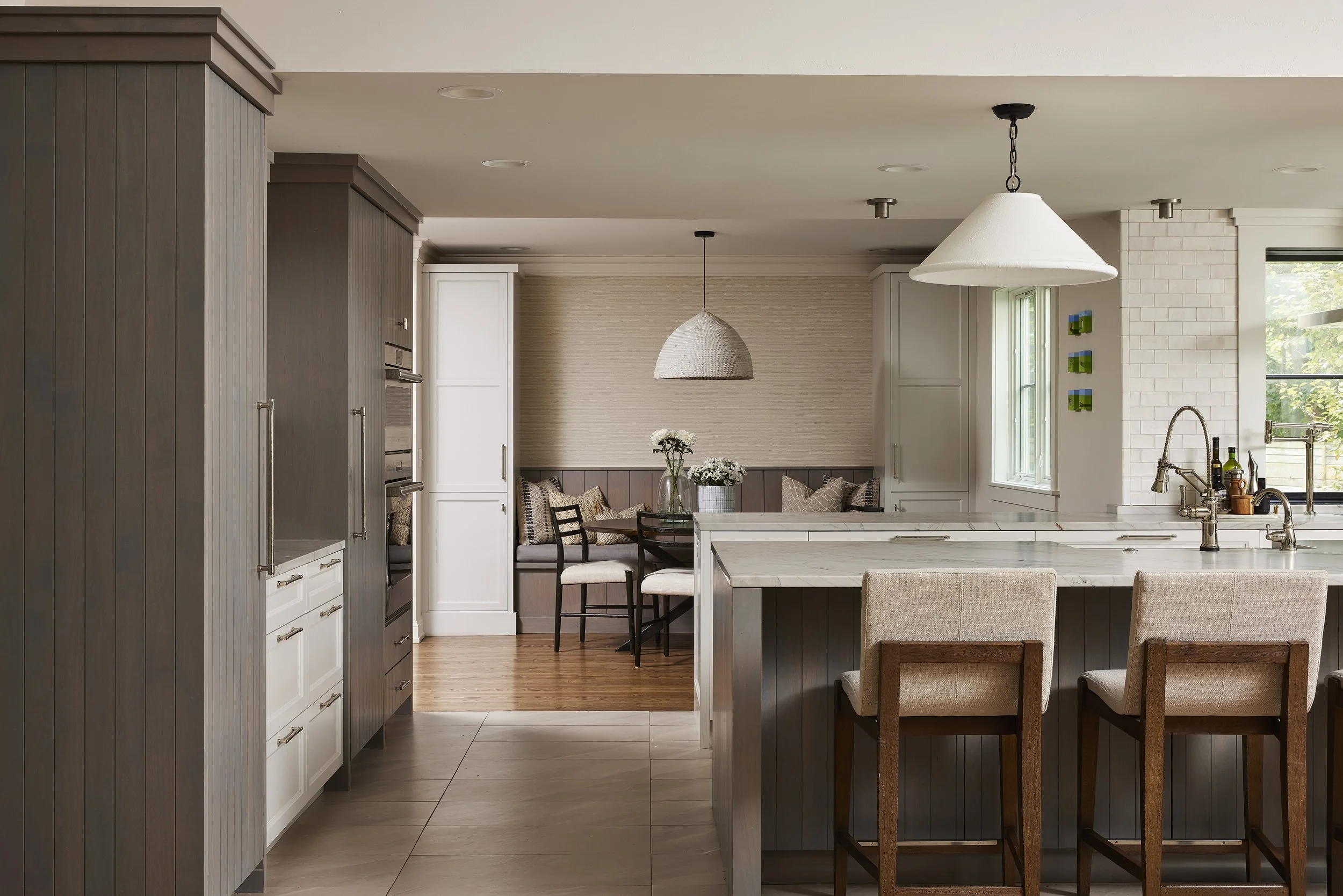 A modern kitchen with a white marble island, beige and brown cabinetry, and a dining nook with a bench and chairs, illuminated by pendant lights, with a window showing greenery.