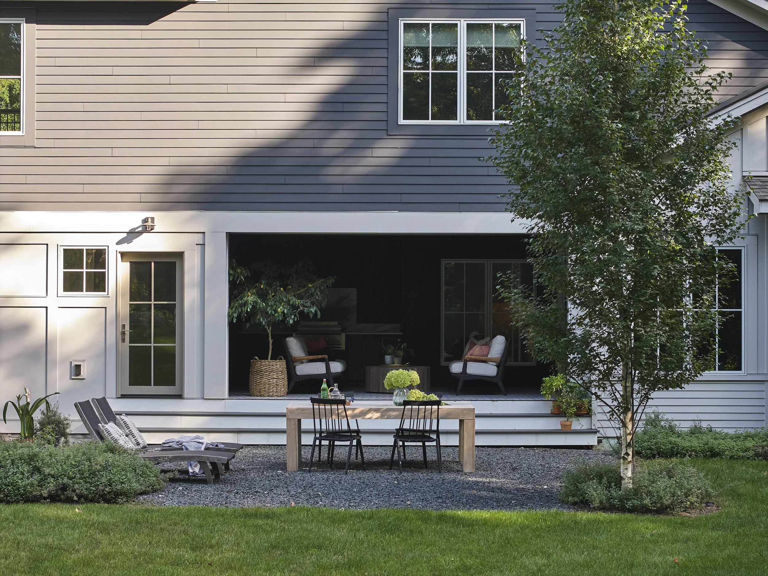Backyard patio with outdoor furniture, a dining table with four chairs, and a lush green lawn with shrubs and a tree.