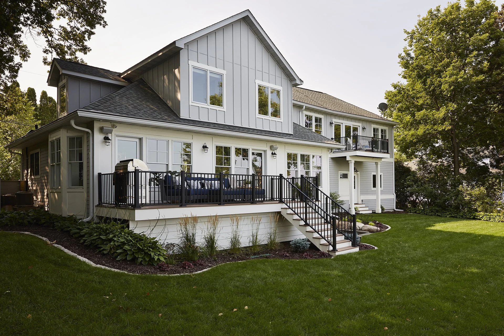 A two-story house with a grey and white exterior, a large deck with black railing, and stairs leading down to a green lawn, surrounded by trees.
