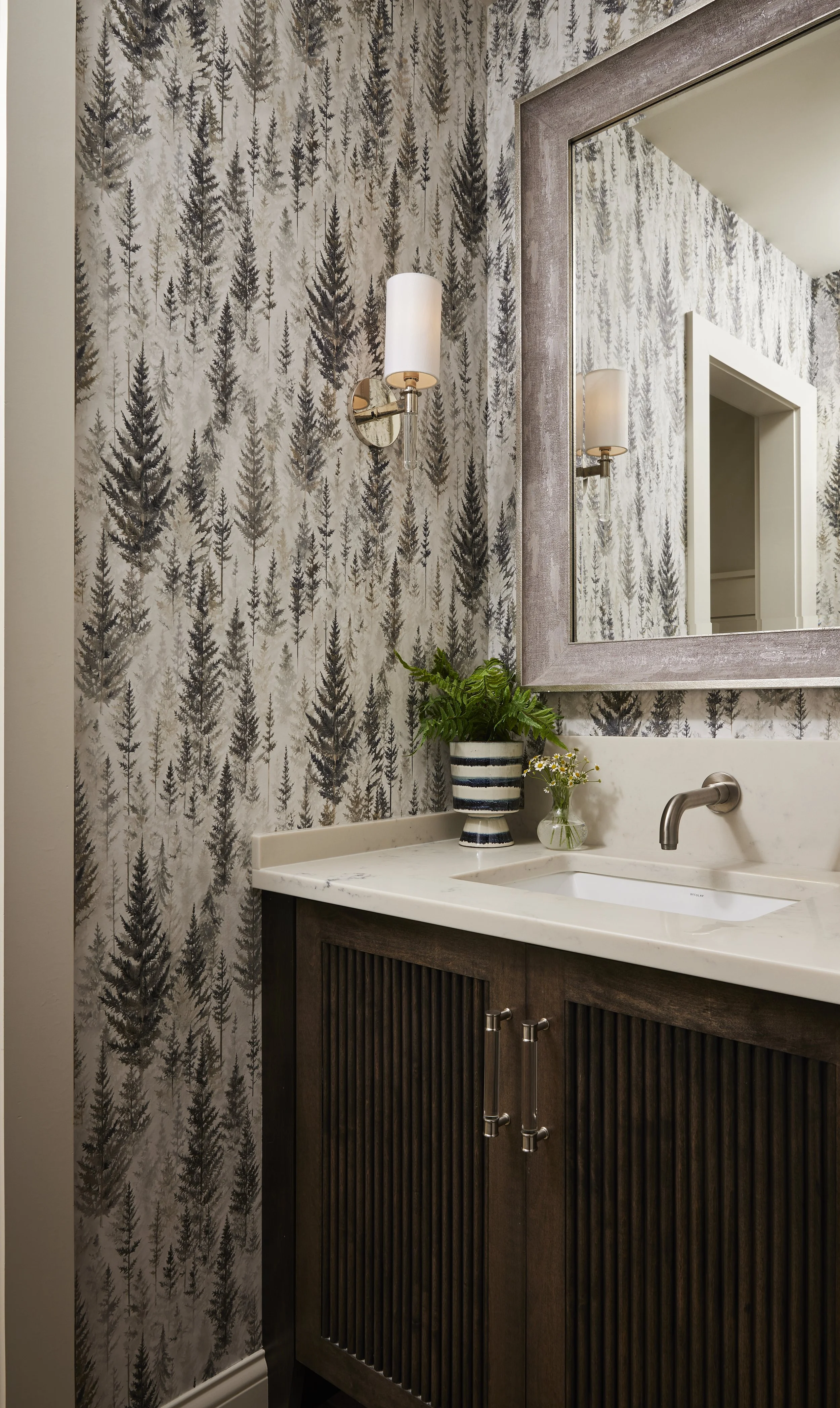 Bathroom vanity with a large mirror, a plant, and a small vase with flowers, illuminated by wall sconces, featuring a wood cabinet and patterned wallpaper with trees.