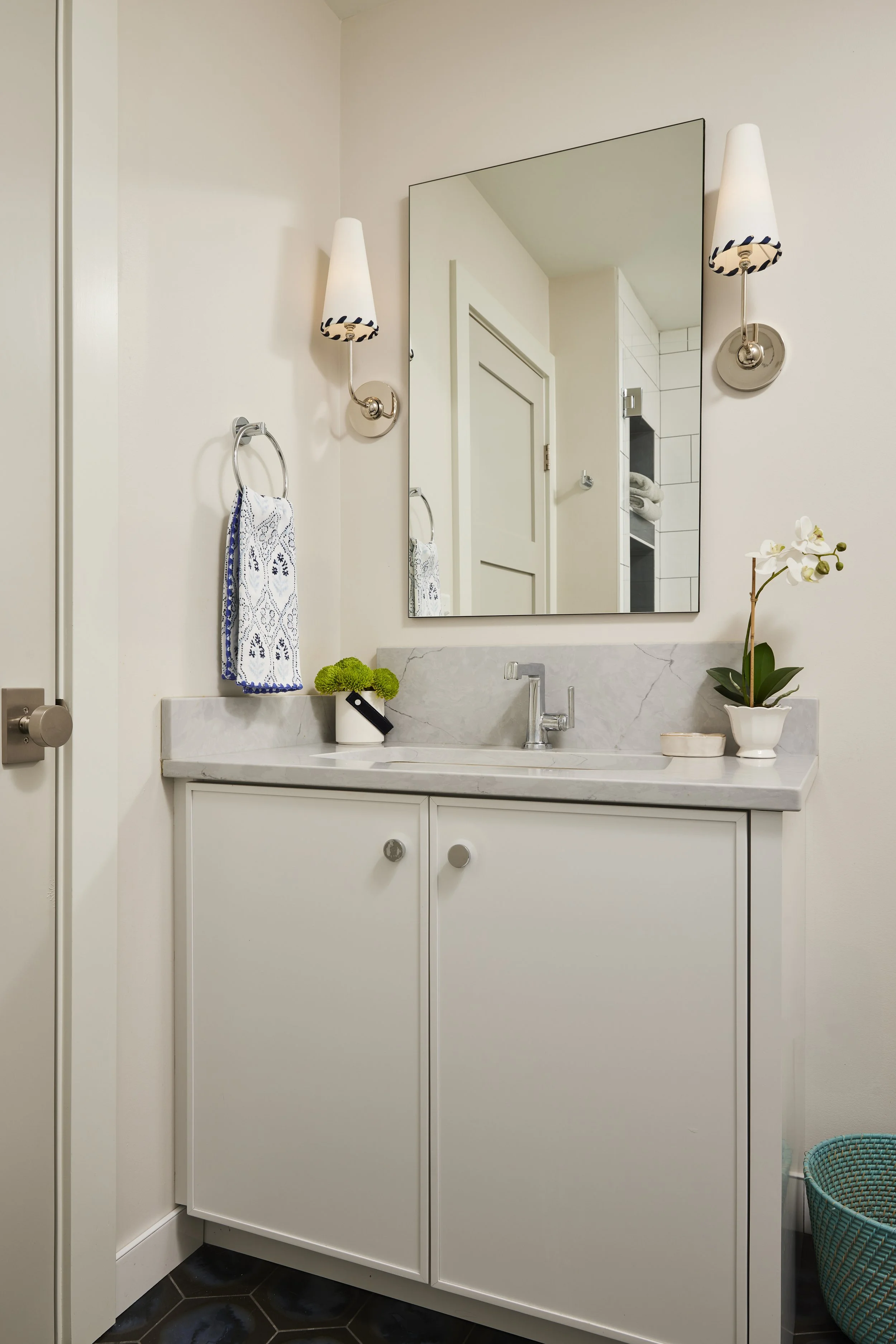 Bathroom with white vanity, marble countertop, and a large mirror. Wall-mounted sconces with white shades flank the mirror. Decor includes a potted orchid and green flowers. A towel ring with a blue and white towel is on the wall. A teal basket is on the floor.