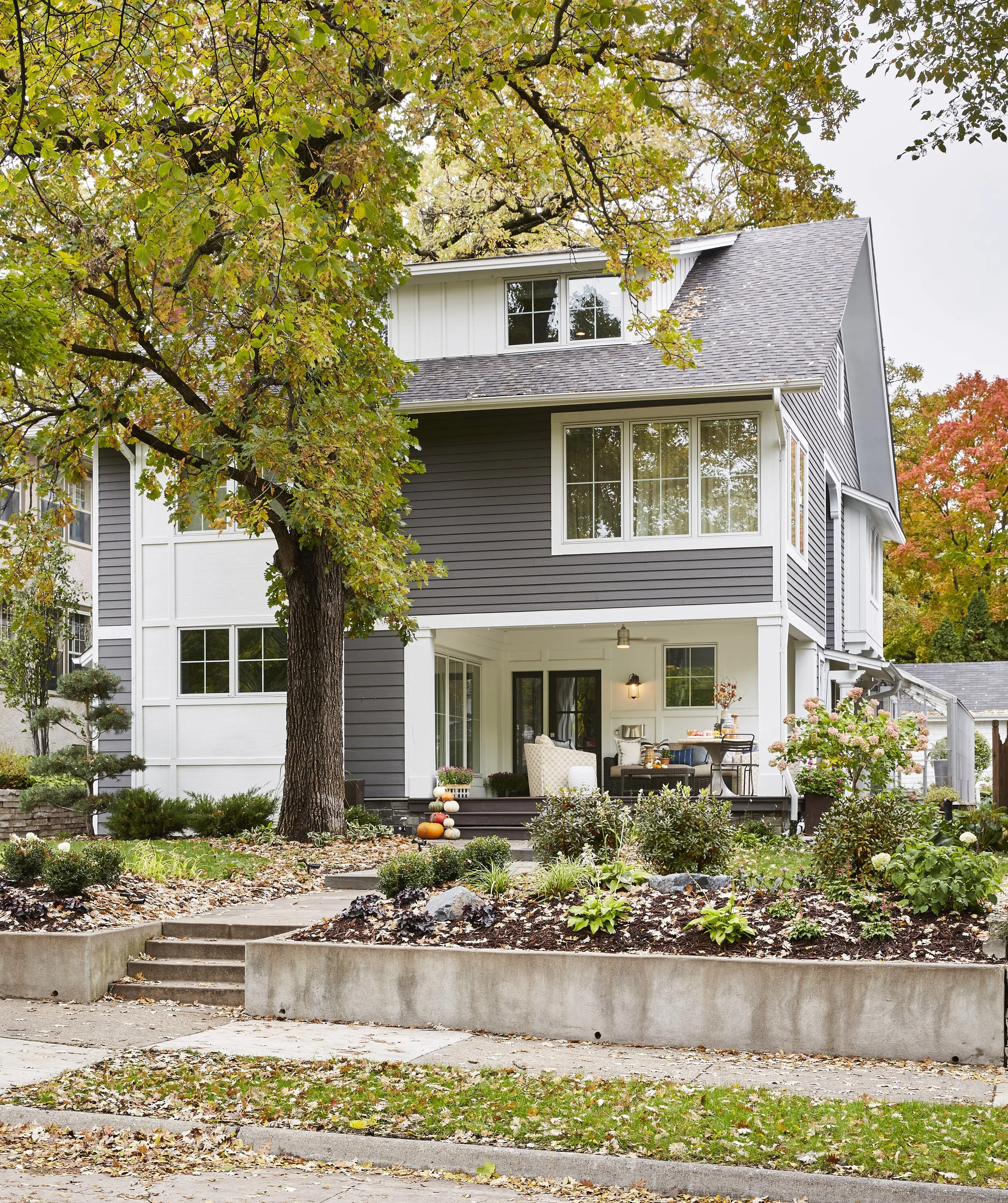A two-story house with gray siding, surrounded by autumn trees and a landscaped yard with plants and flowers. There's a porch with outdoor furniture and fall decorations.