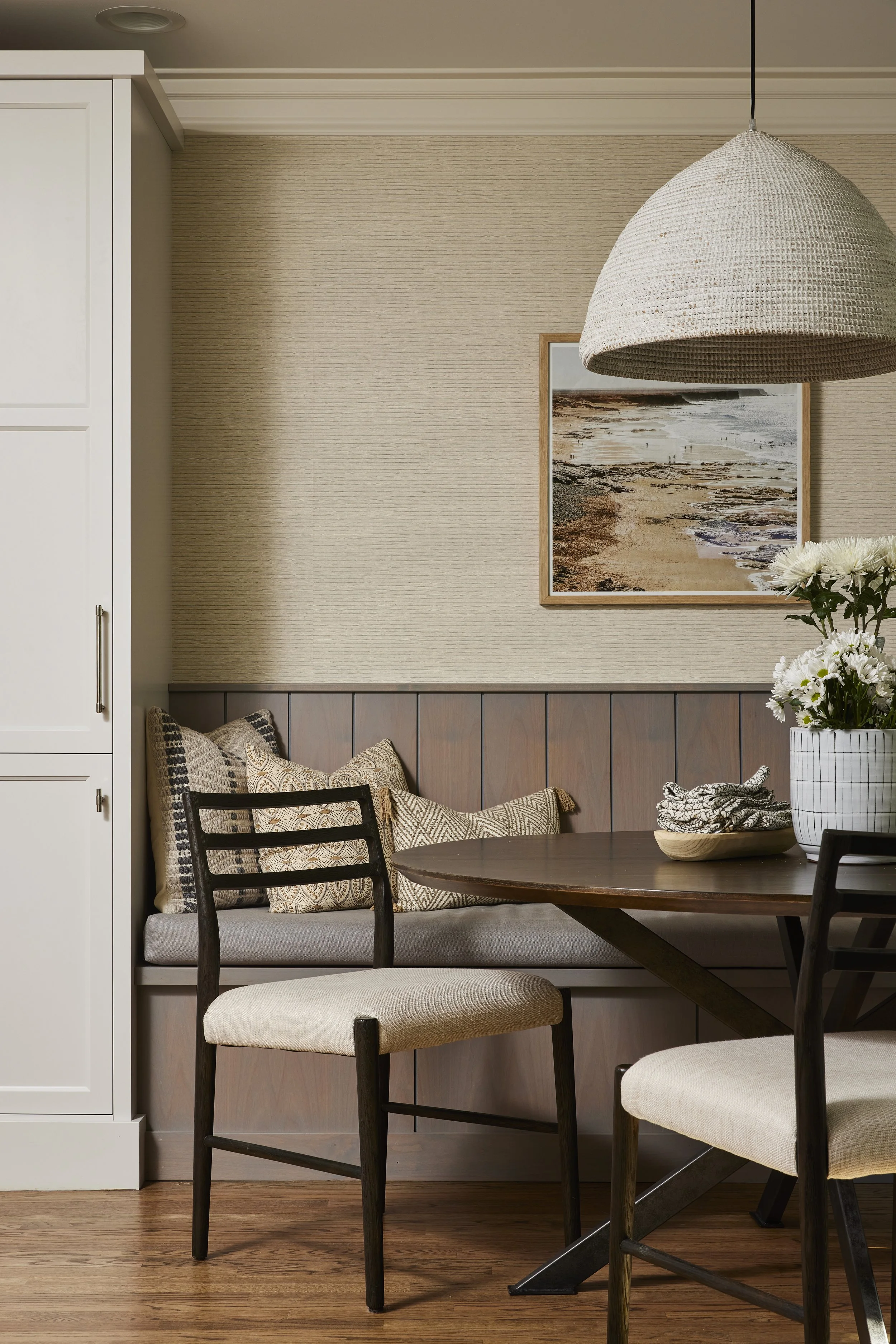 A cozy dining nook with a wooden table, cushioned bench with decorative pillows, surrounded by two upholstered chairs with dark wooden frames. A large hanging woven light fixture, a flower vase with white flowers, and a framed beach photograph on a beige textured wall complete the scene.