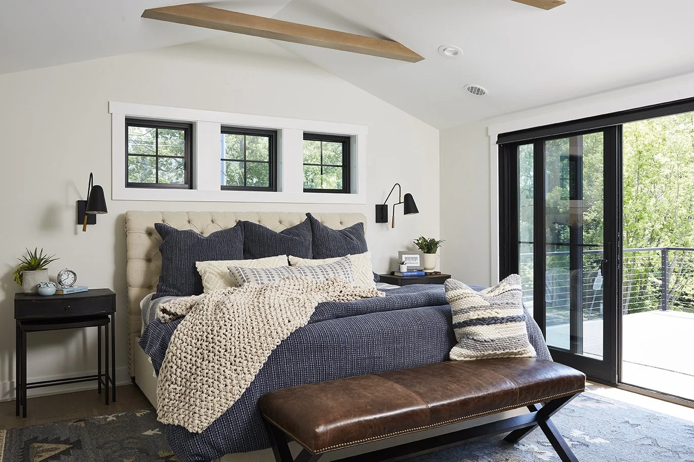 A cozy bedroom with a tufted beige headboard, dark blue bedding, and multiple pillows. There are black wall-mounted reading lamps, black side tables with decor, an outdoor balcony visible through large sliding glass doors, and a brown leather bench at the foot of the bed.