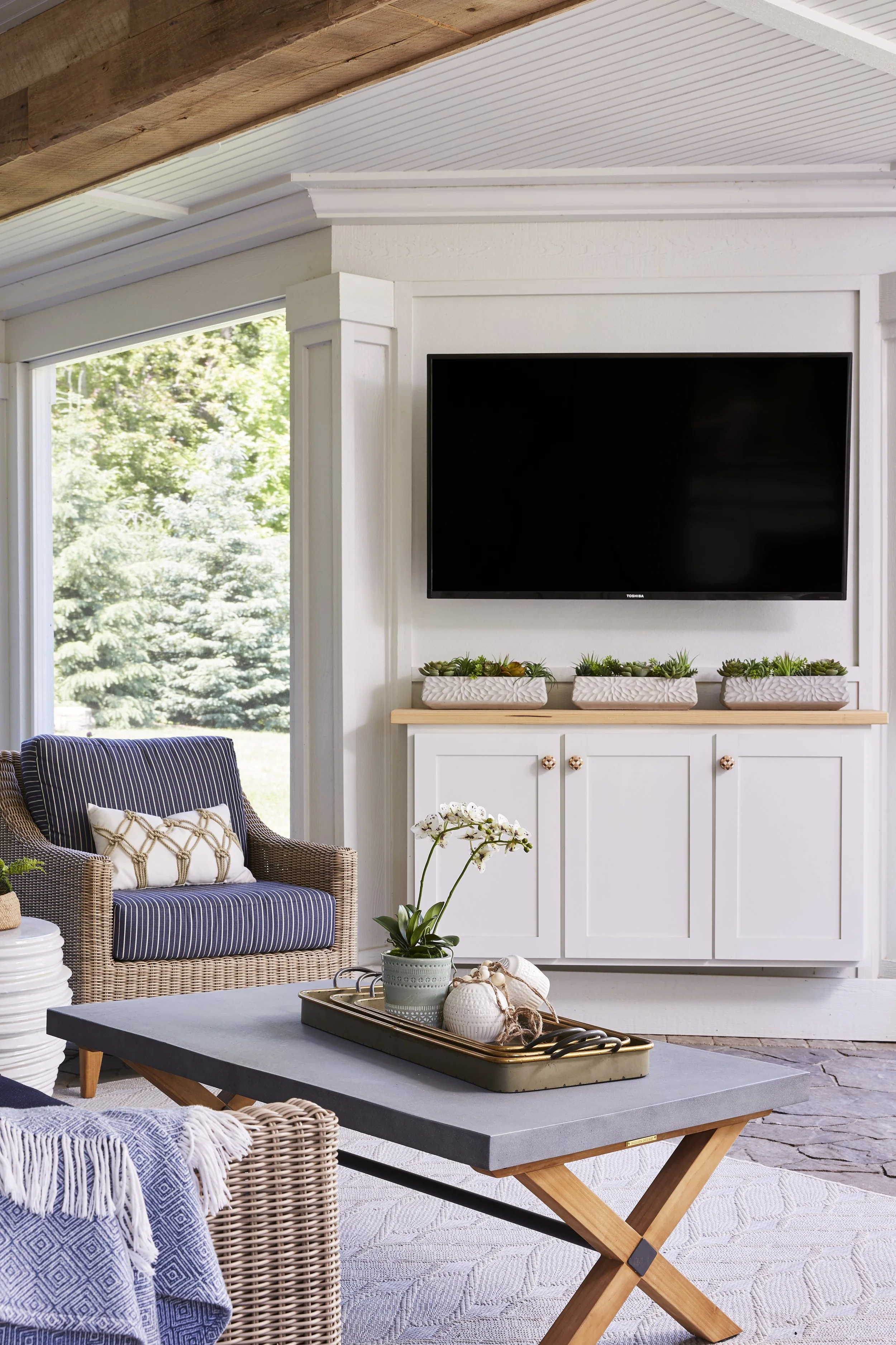 Living room with a wall-mounted flat-screen TV, a wicker armchair with a patterned pillow, a coffee table with a flower vase, and a cabinet with potted succulents, next to a large window showing greenery outside.