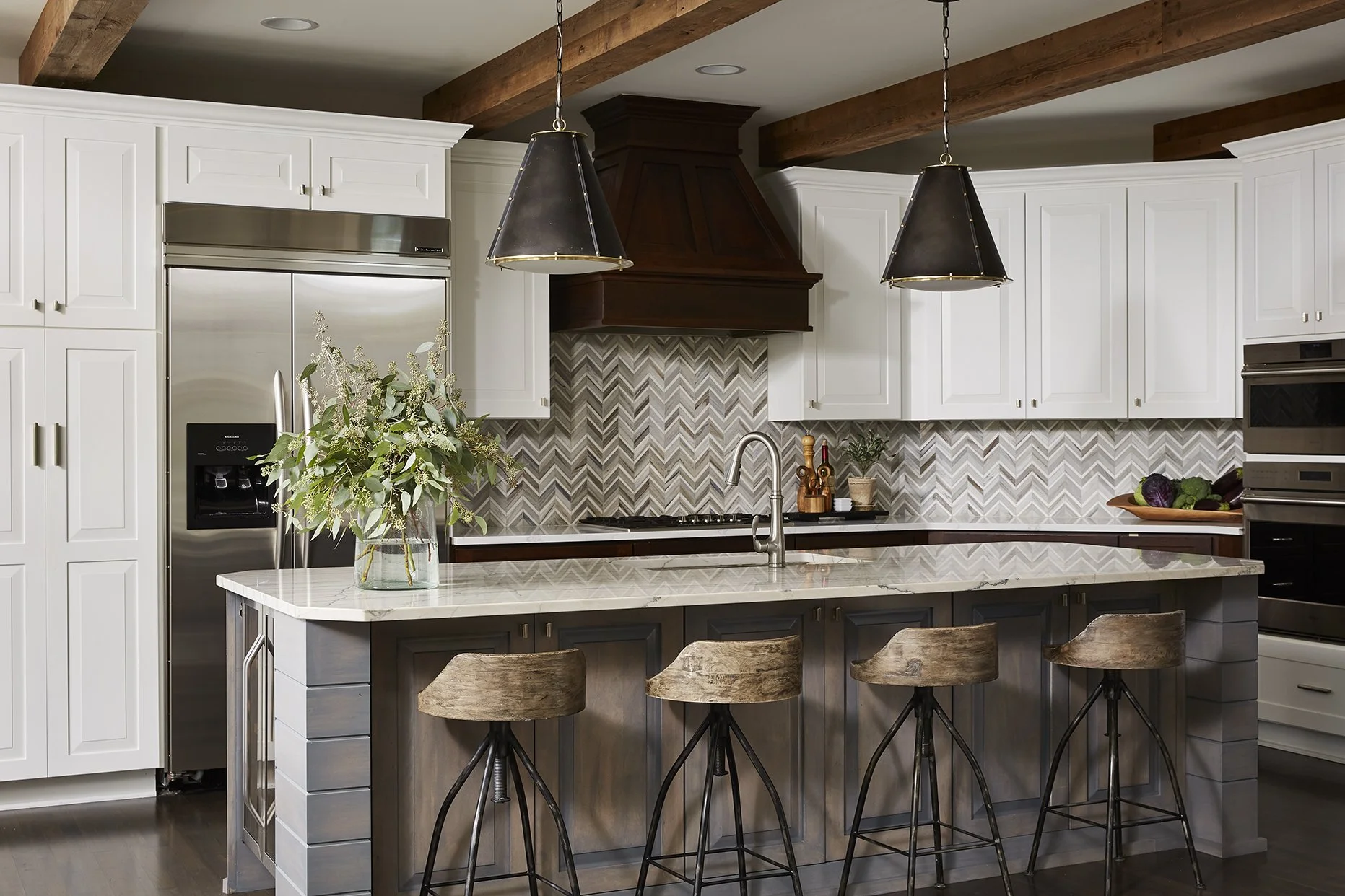 Modern kitchen with white cabinetry, wooden beams, stainless steel appliances, a marble island with four wooden and metal barstools, pendant lights, a vase with greenery, and a patterned backsplash.