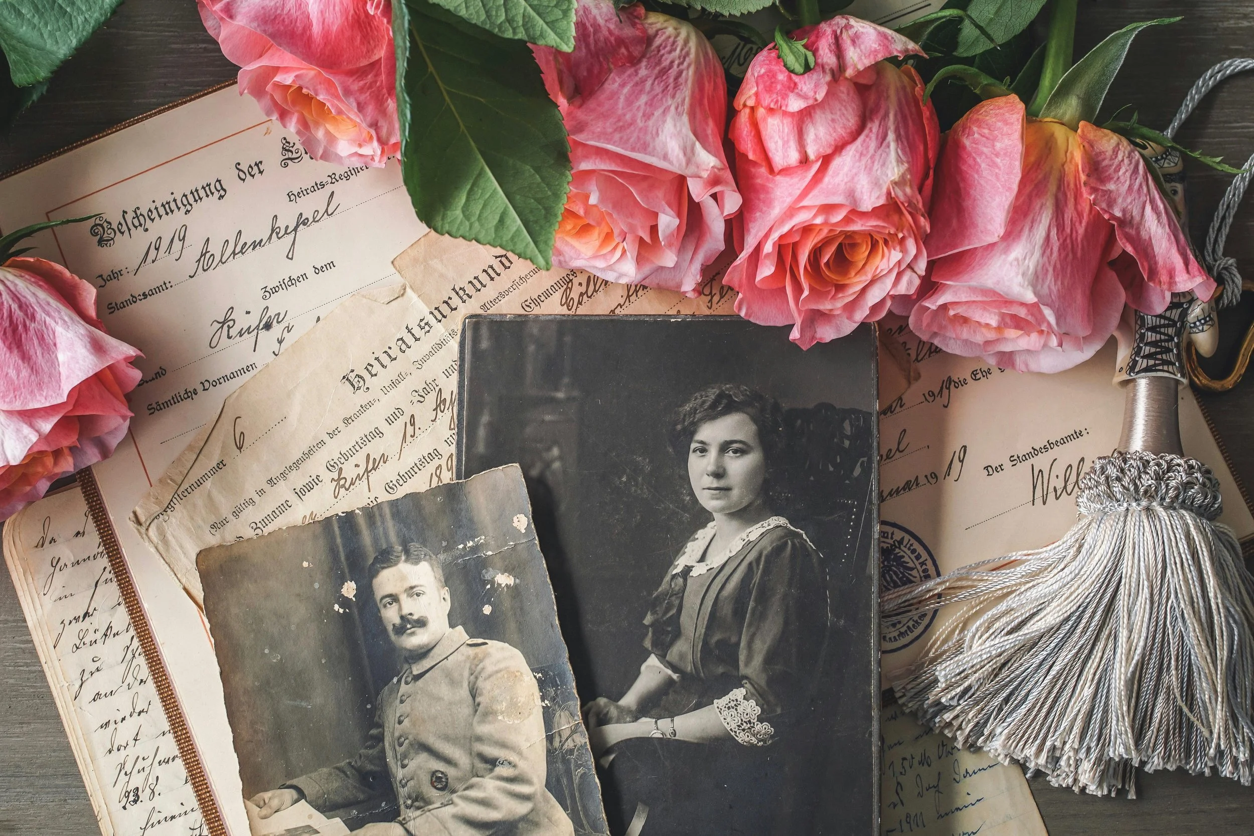 A collection of vintage photographs, old documents in German, pink roses, and a decorative sword with a tassel.