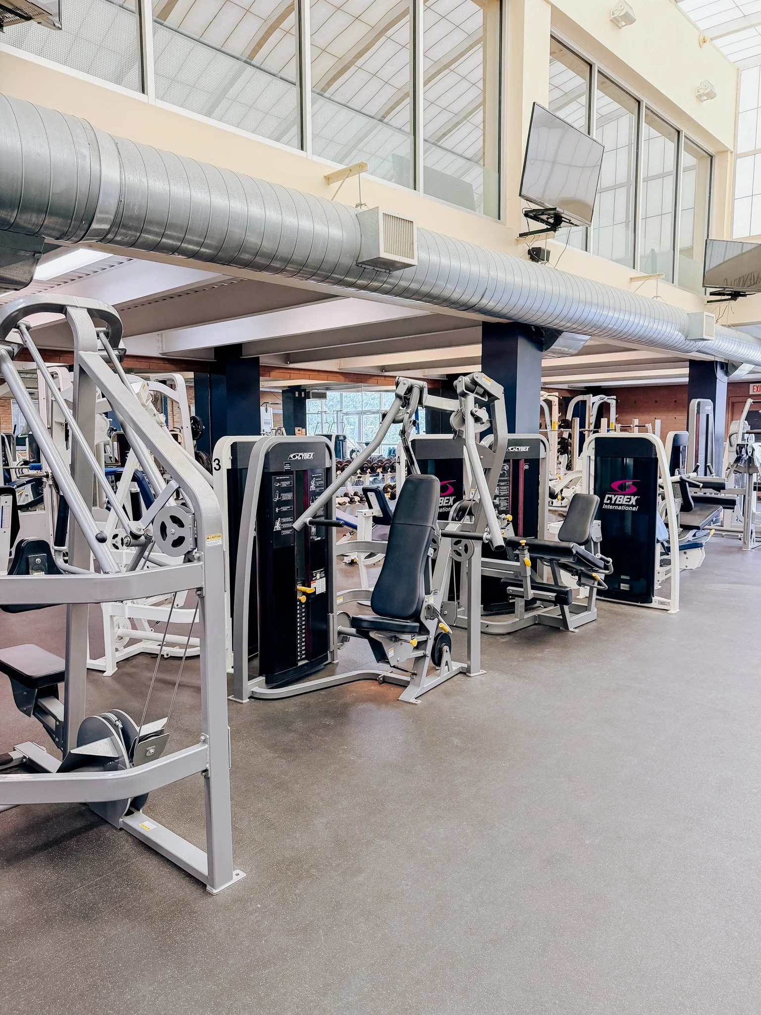 A gym interior featuring various exercise equipment including weight machines and cardio equipment in a spacious, well-lit area with large windows and ceiling-mounted displays.