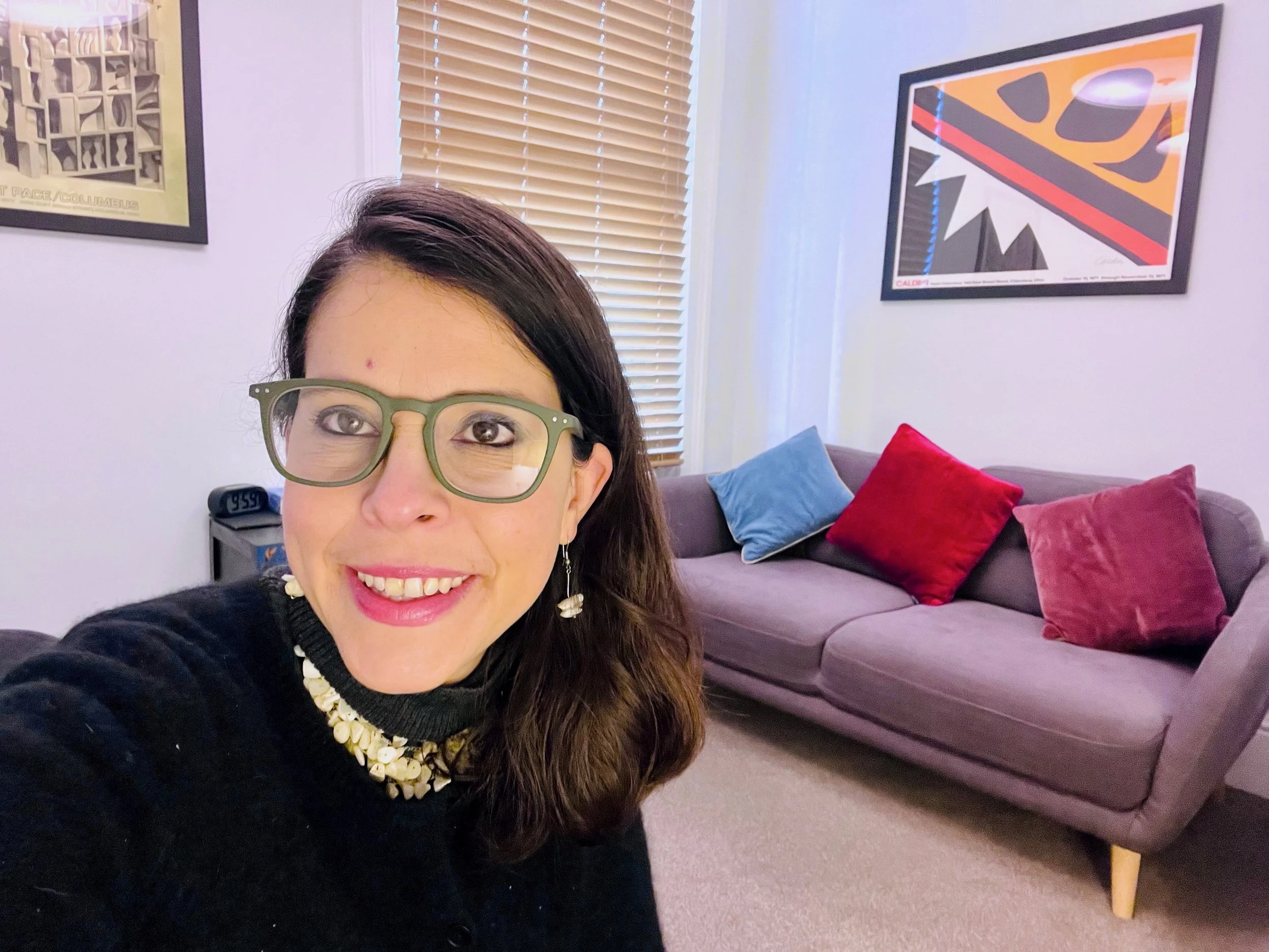 A woman with long brown hair, glasses, and earrings taking a selfie in a living room with a purple sofa decorated with red, pink, and blue cushions, framed artwork on the walls, and wooden blinds covering a window.