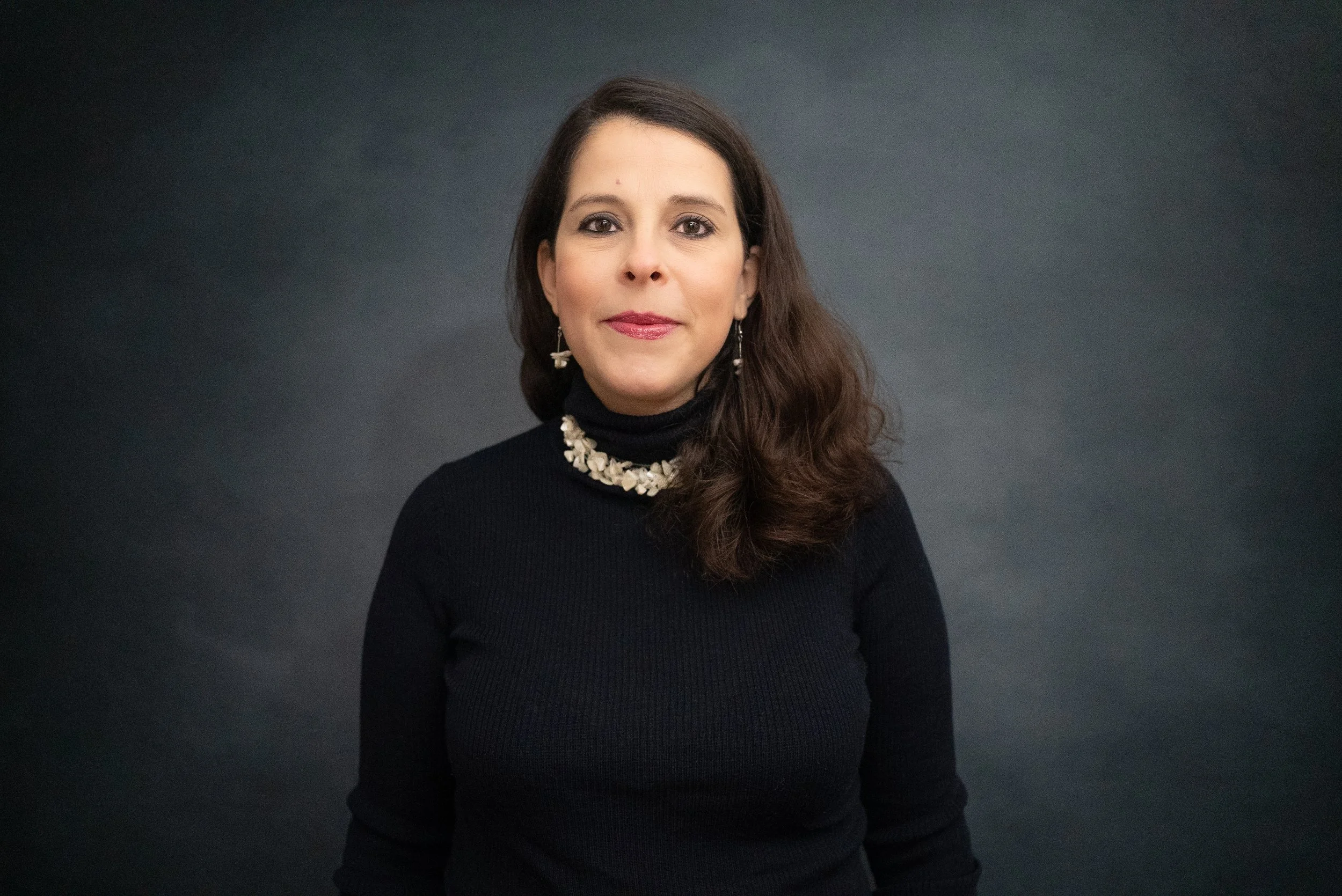 A woman with long brown hair wearing a black turtleneck and pearl jewelry, posing against a dark gray background.