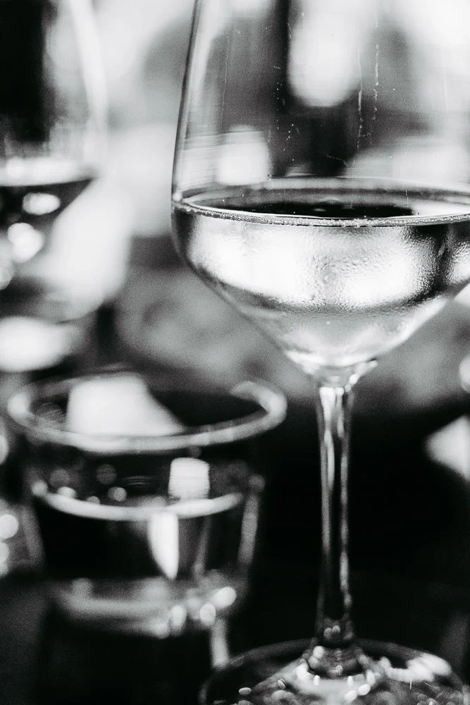 Weißweinglas mit Wasser auf einem dunklen Tisch, im Hintergrund Gläser und Dekorationen, schwarz-weiß Foto.