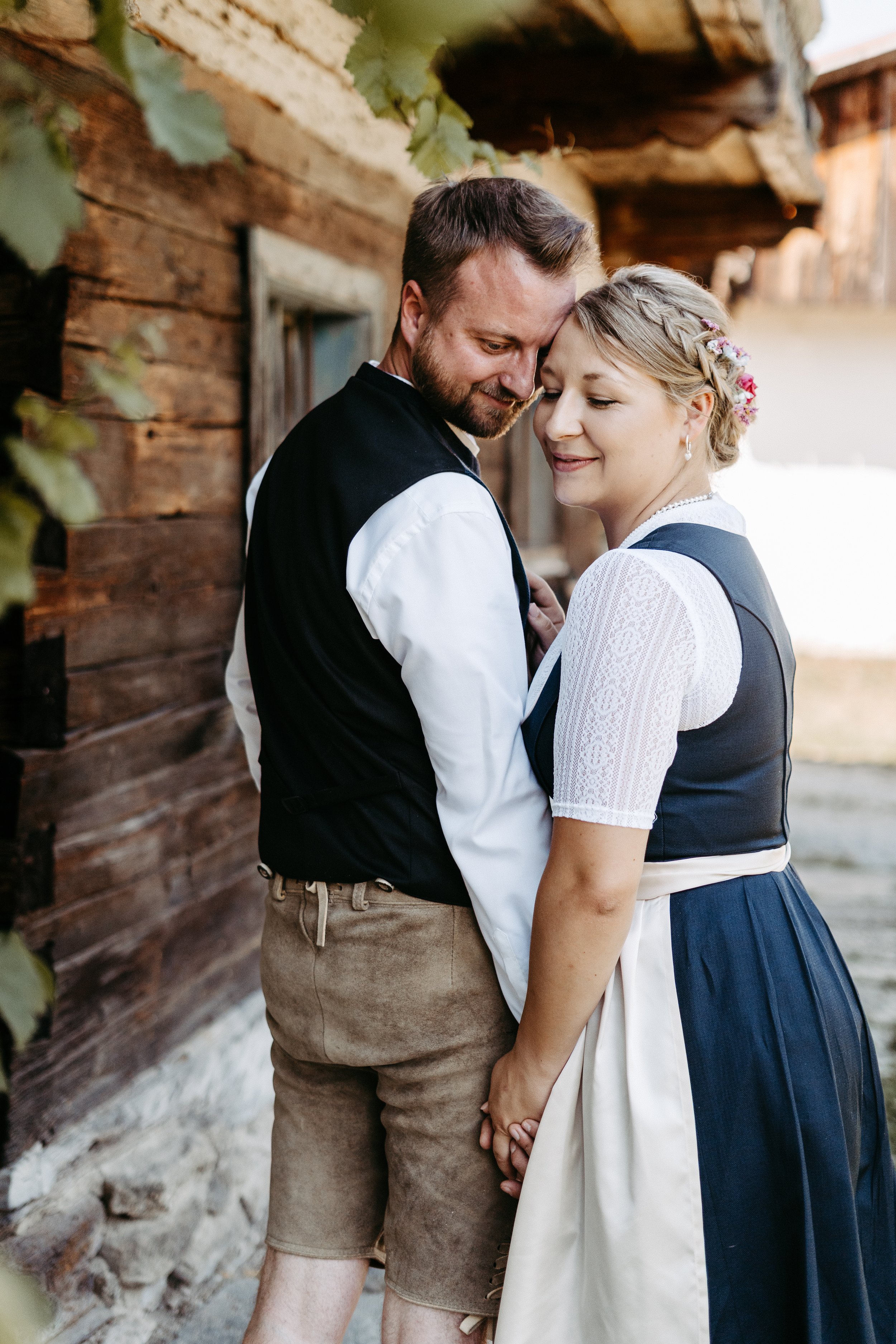Ein glückliches Paar in traditioneller Kleidung steht vor einem Holzhaus, sie berühren sich zärtlich und lächeln.