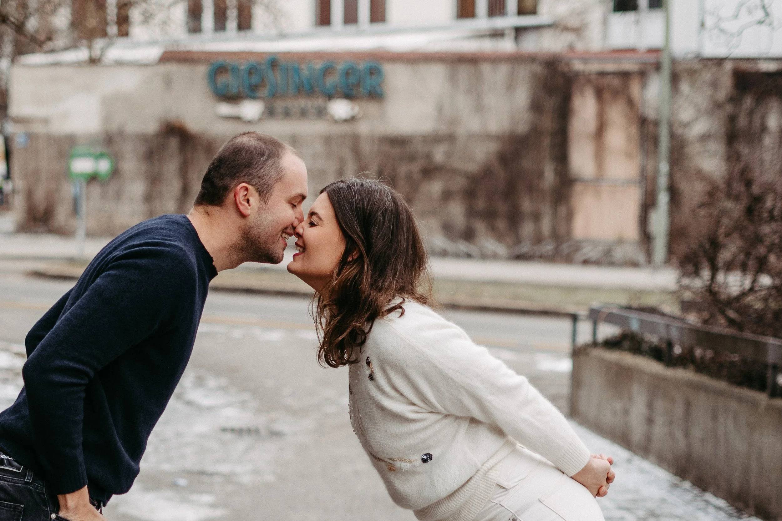 Paar, das sich in der Öffentlichkeit vor einer Wand mit der Aufschrift 'GIESINGER' gegenübersteht, lächelnd und die Nasen berührend