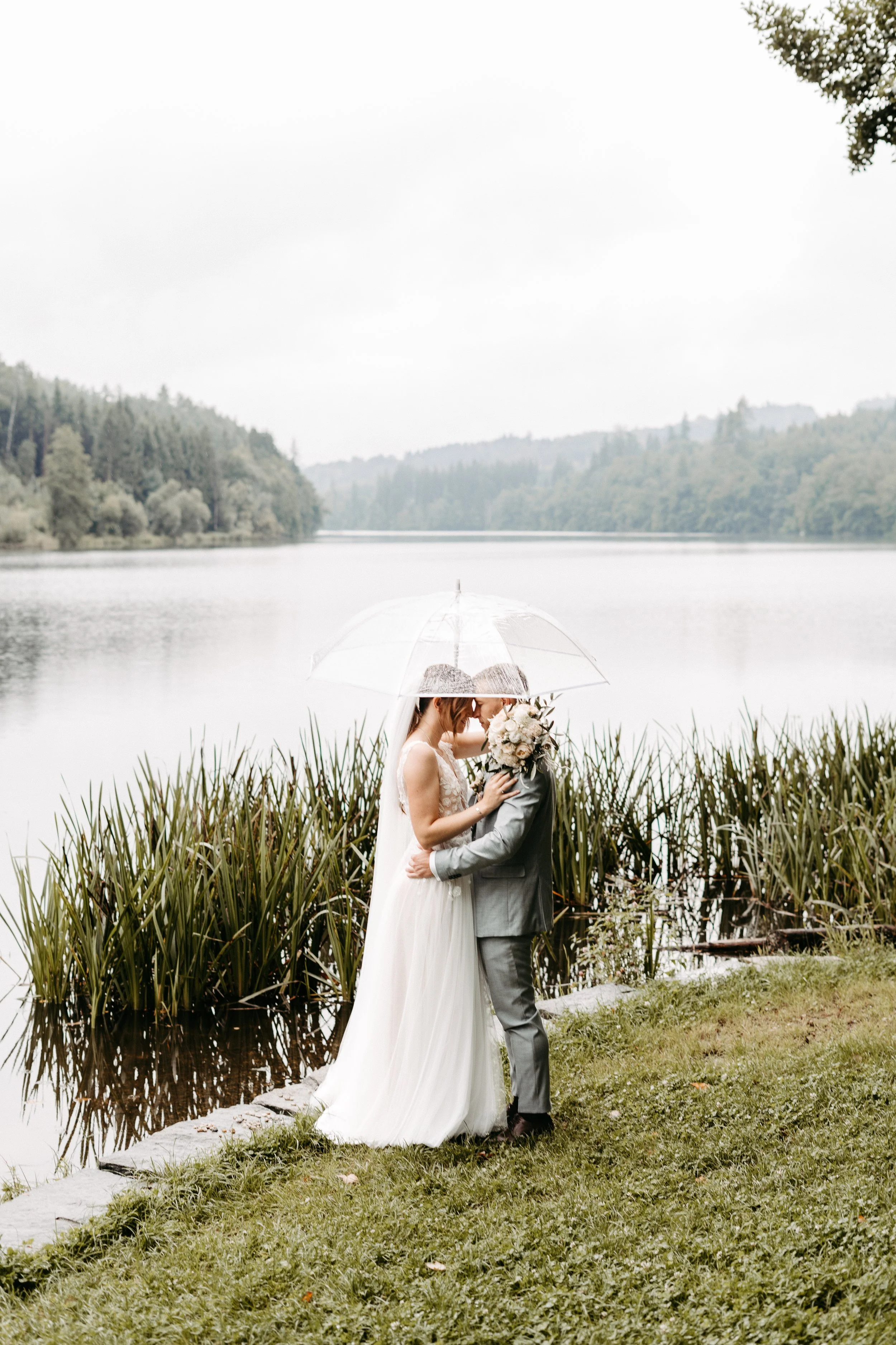 Ein Brautpaar steht sich an einem See mit Regenschirm umarmend und küsst, während es regnet, umgeben von Büschen und Bäumen.