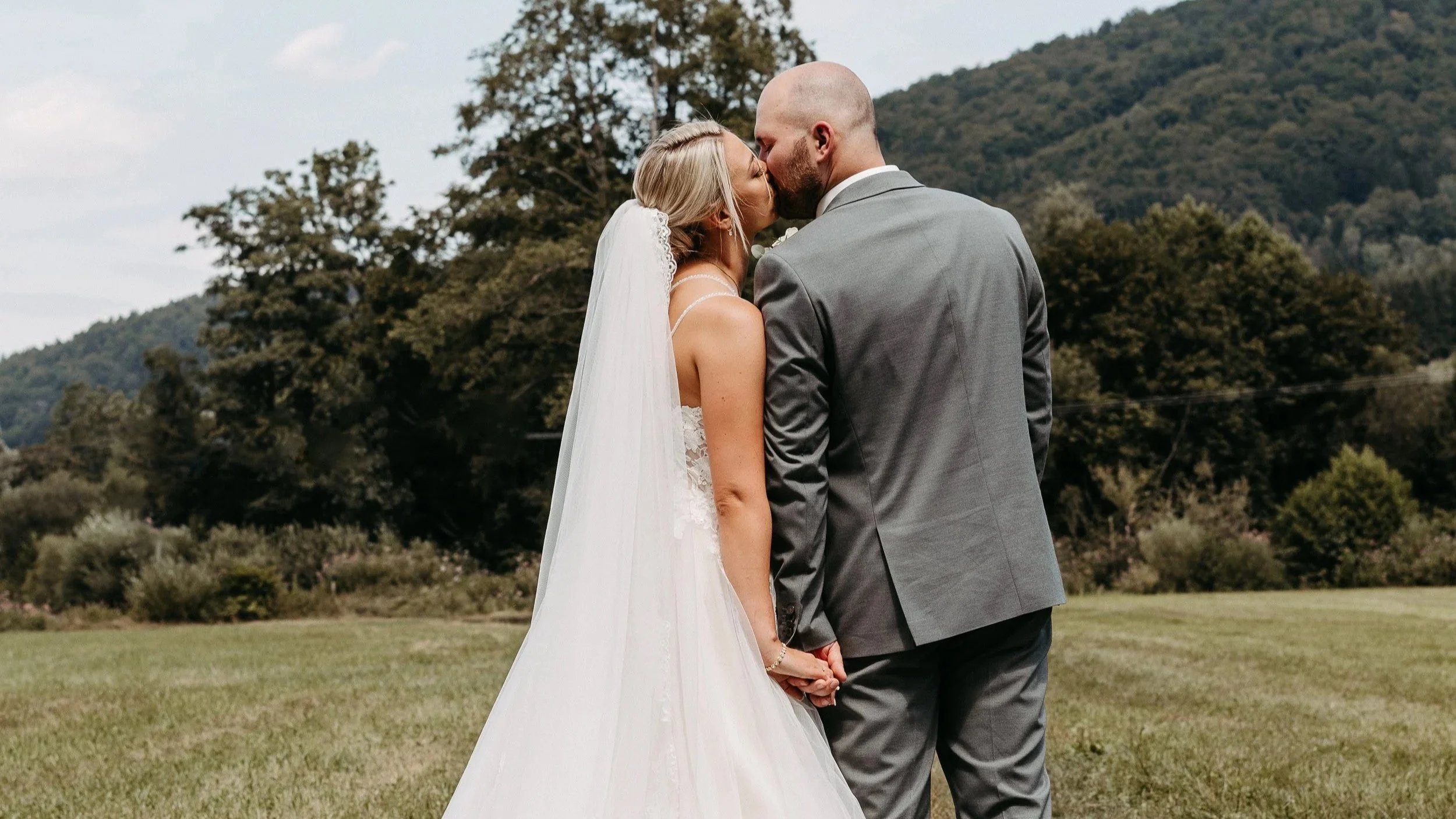 Ein Brautpaar küsst sich draußen vor einer grünen Landschaft mit Bäumen und Bergen im Hintergrund.