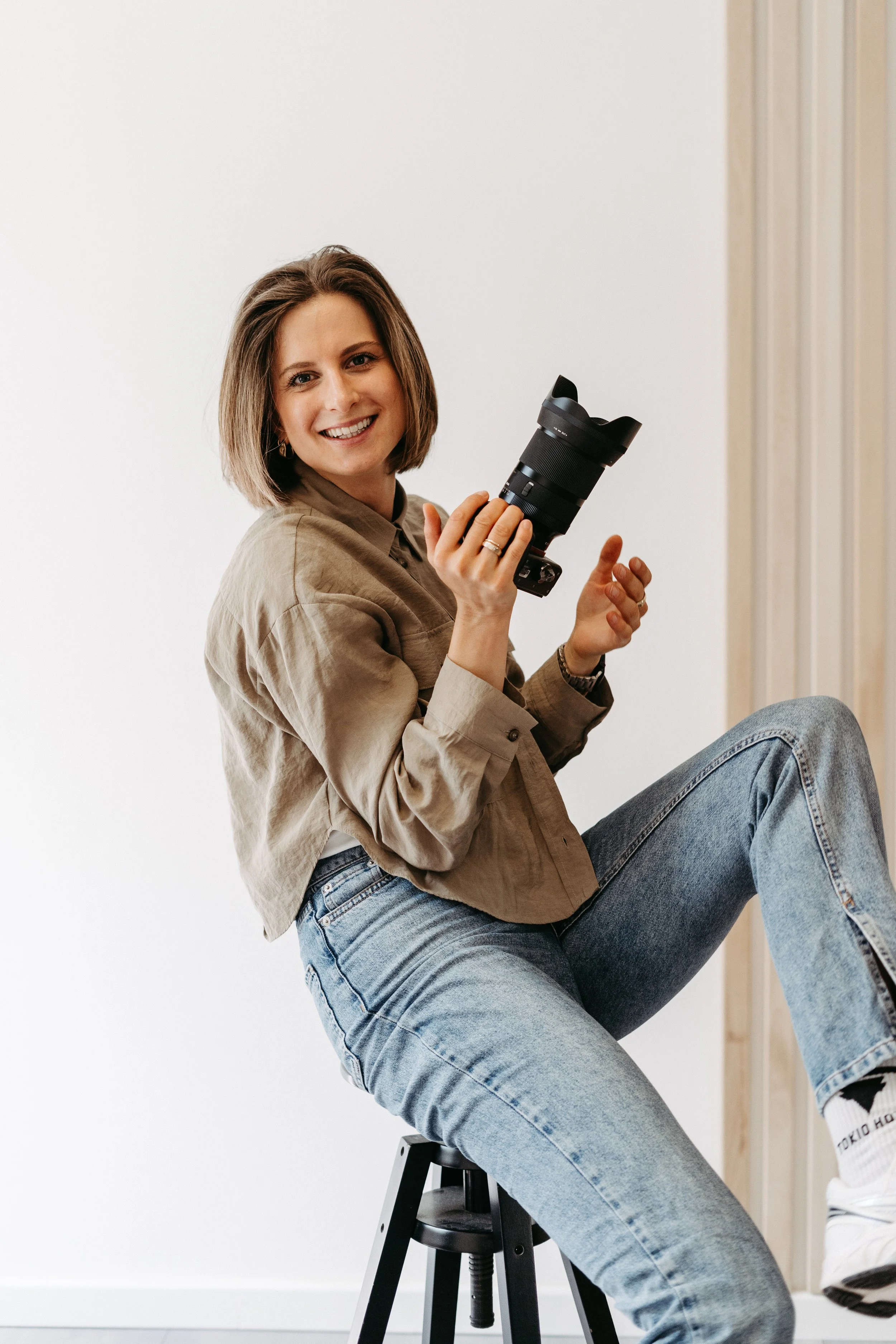 Eine lächelnde junge Frau sitzt auf einem Hocker, hält eine Kamera und trägt einen beigen Blazer und Blue Jeans.