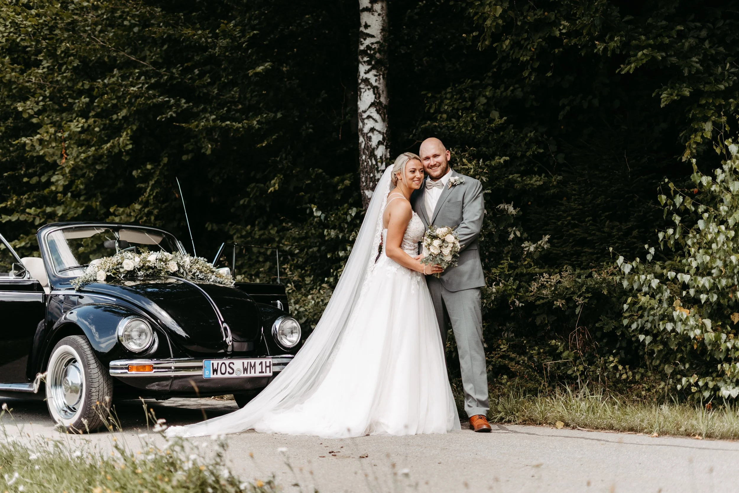Ein lächelndes Brautpaar steht vor einem schwarzen Oldtimer, der mit Blumen geschmückt ist, im Freien vor einem dunklen grünen Waldrand.
