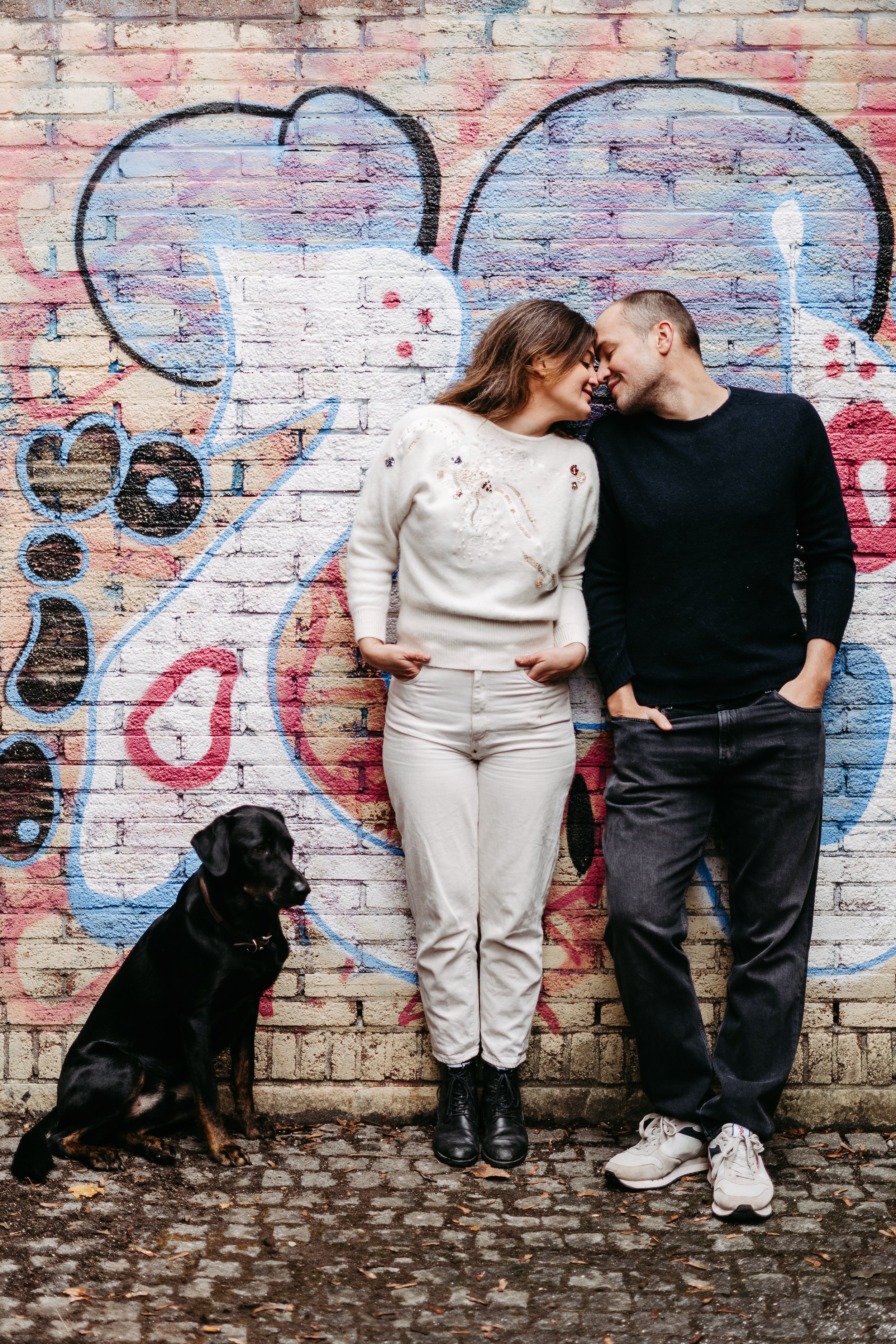 Ein Paar steht vor einer mit Graffiti bemalten Ziegelwand, die ein großes Herz zeigt. Der Hund sitzt auf dem Kopfsteinpflaster vor ihnen.