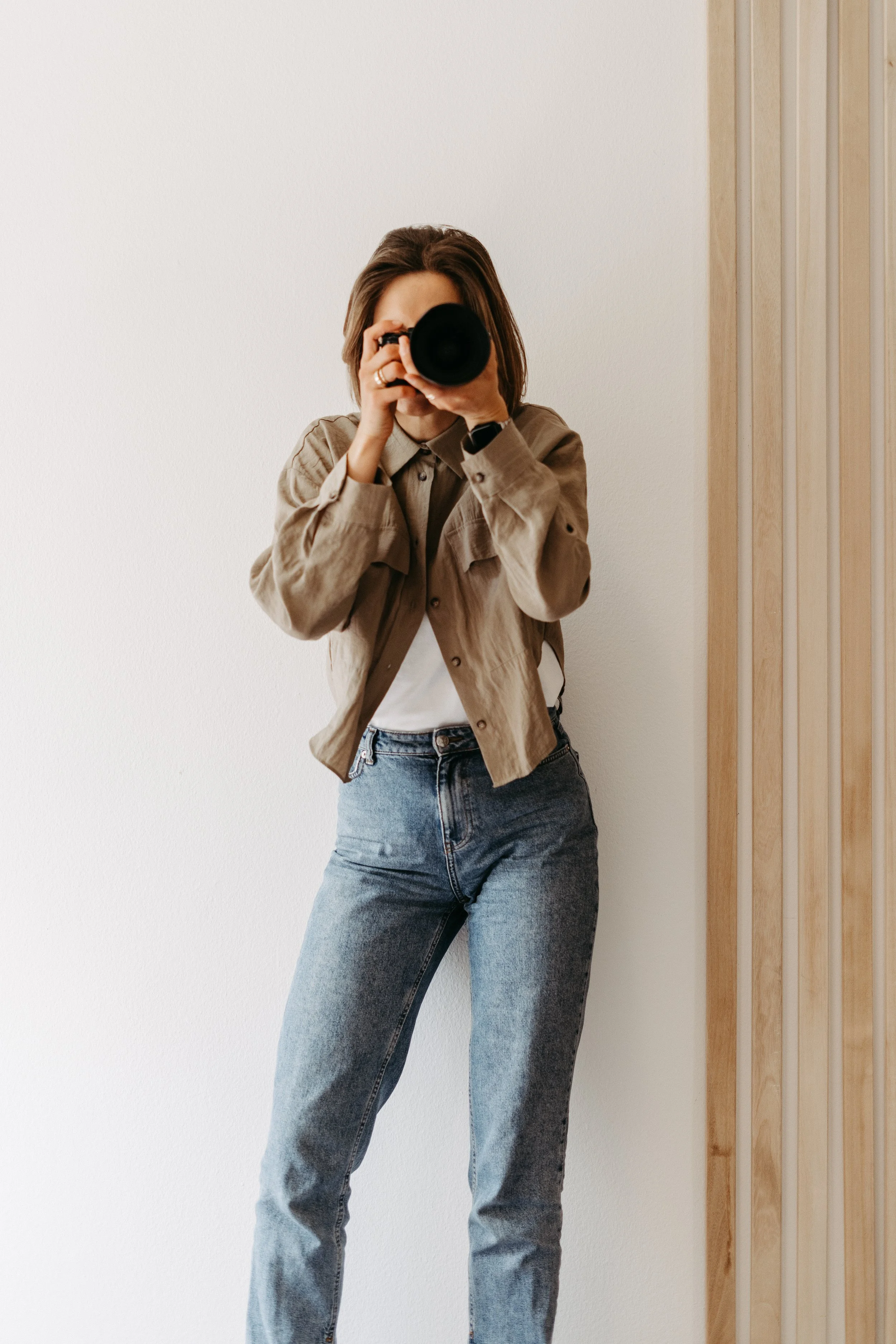 Frau macht ein Foto mit einer Kamera, stehend vor weißer Wand und Holzverkleidung.