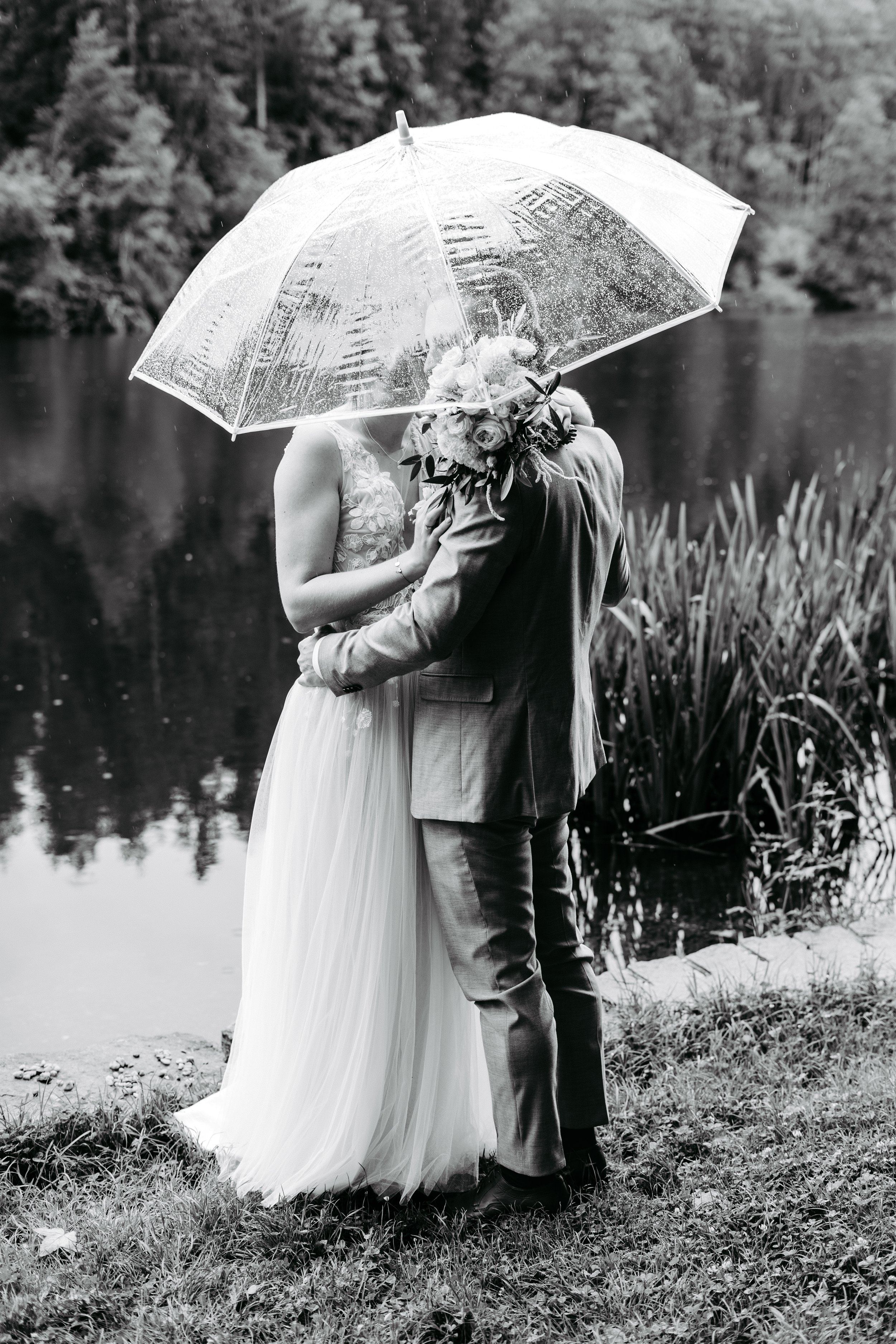 Ein Paar küssend unter einem transparenten Regenschirm an einem Teich mit Bäumen im Hintergrund, im Schwarz-Weiß-Format.