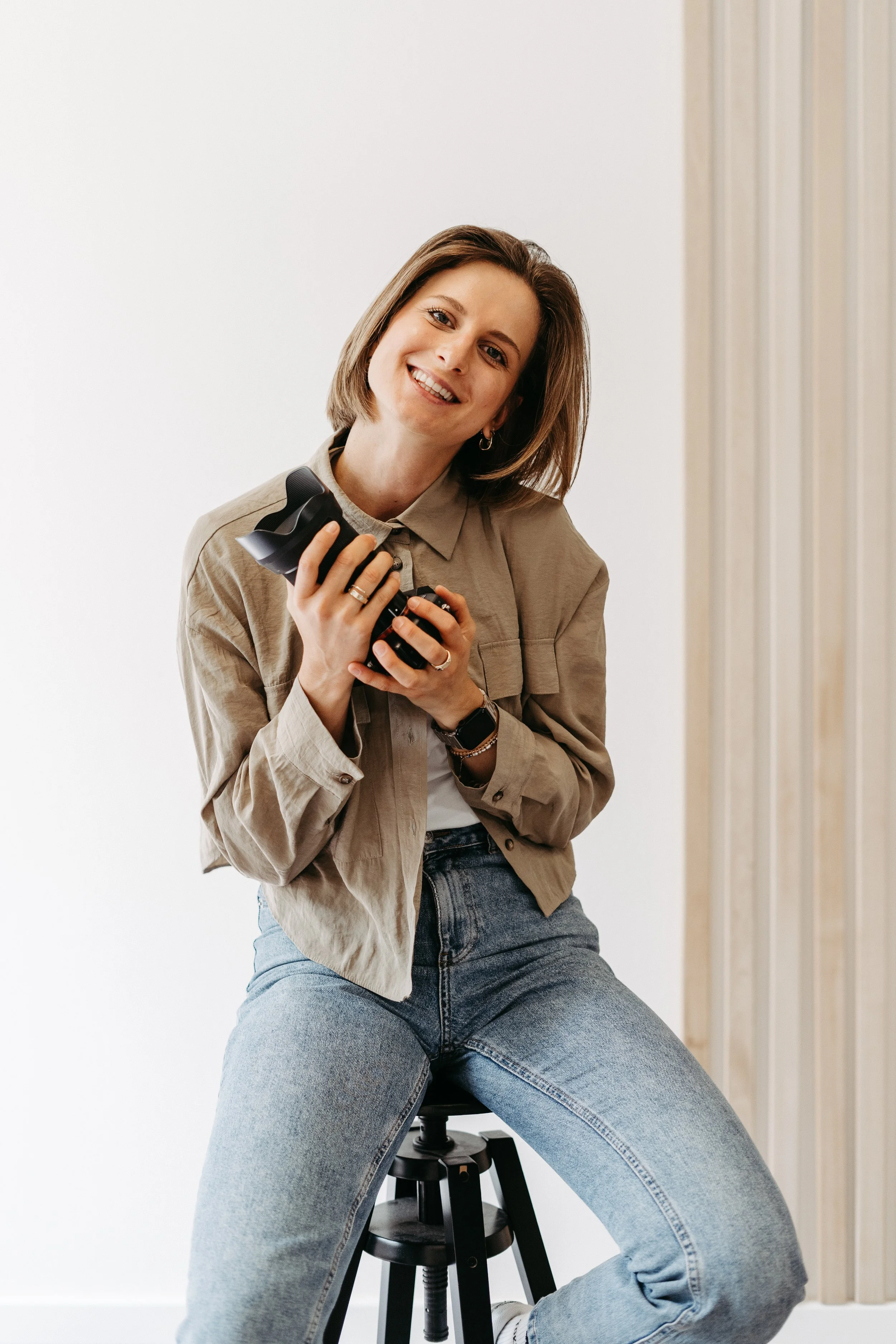 Junge Frau sitzt auf einem Hocker, hält eine Kamera und lächelt in die Kamera, trägt eine beige Jacke und blaue Jeans, im Hintergrund sind weiße Wände und eine Holzverkleidung.