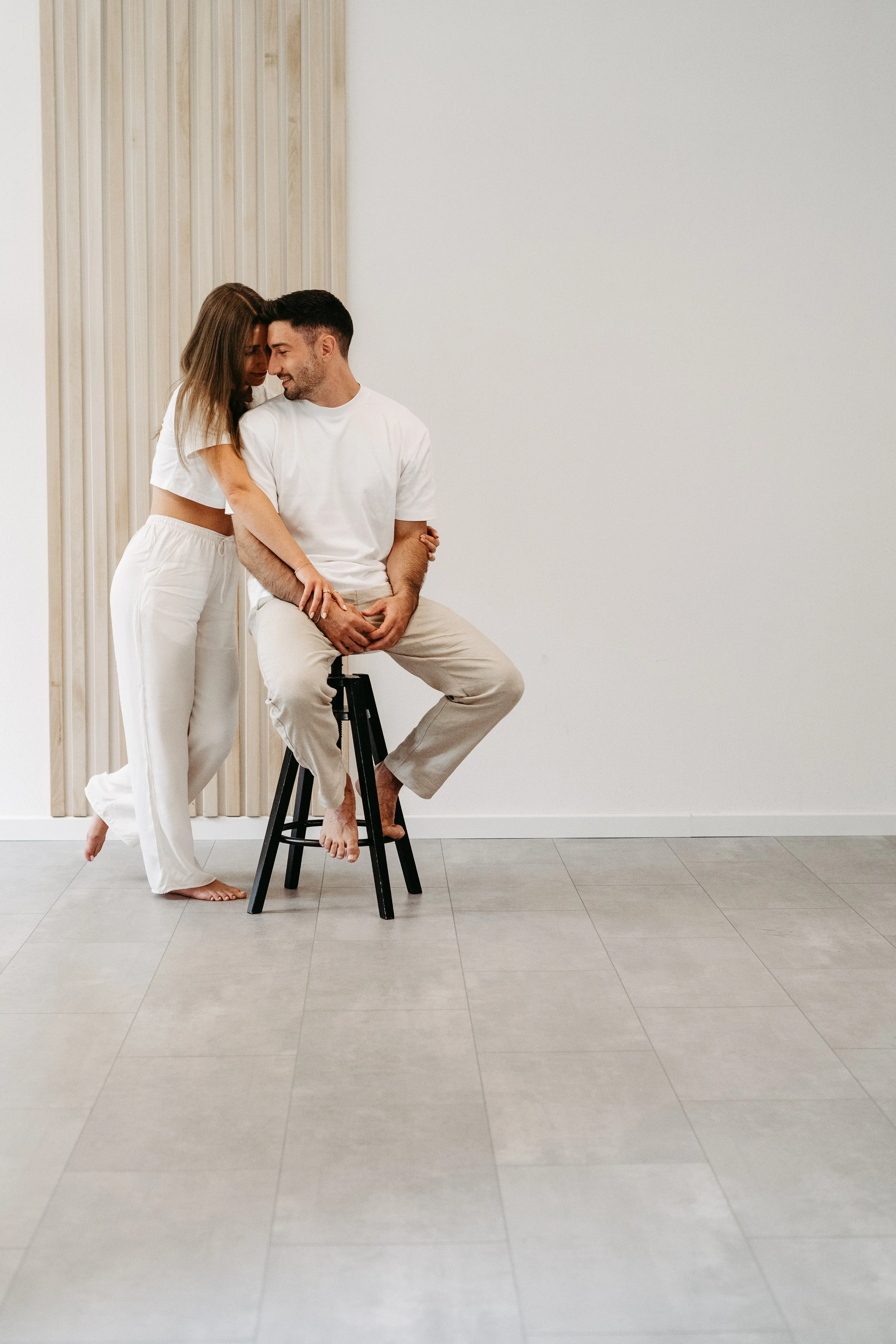 Ein Paar sitzt auf einem Barhocker in einem minimalistischen Raum, umarmt sich liebevoll und lacht.