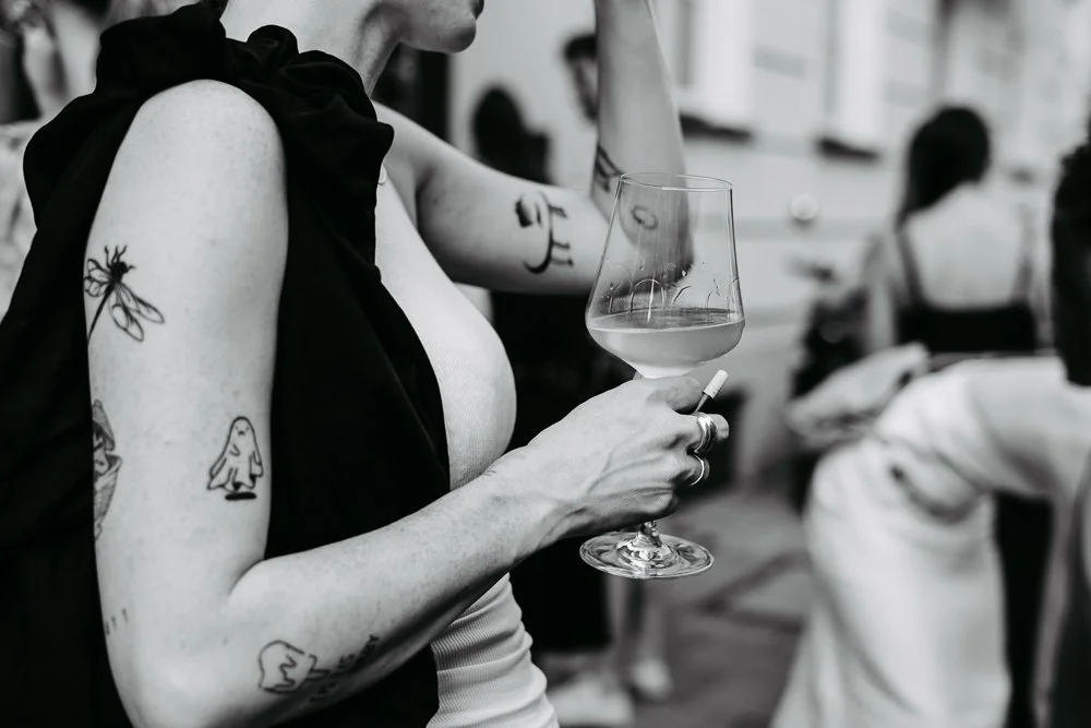 Nahaufnahme einer Frau mit Tätowierungen, die ein Weinglas hält, im Hintergrund andere Personen in einem angesagten Café oder Restaurant.